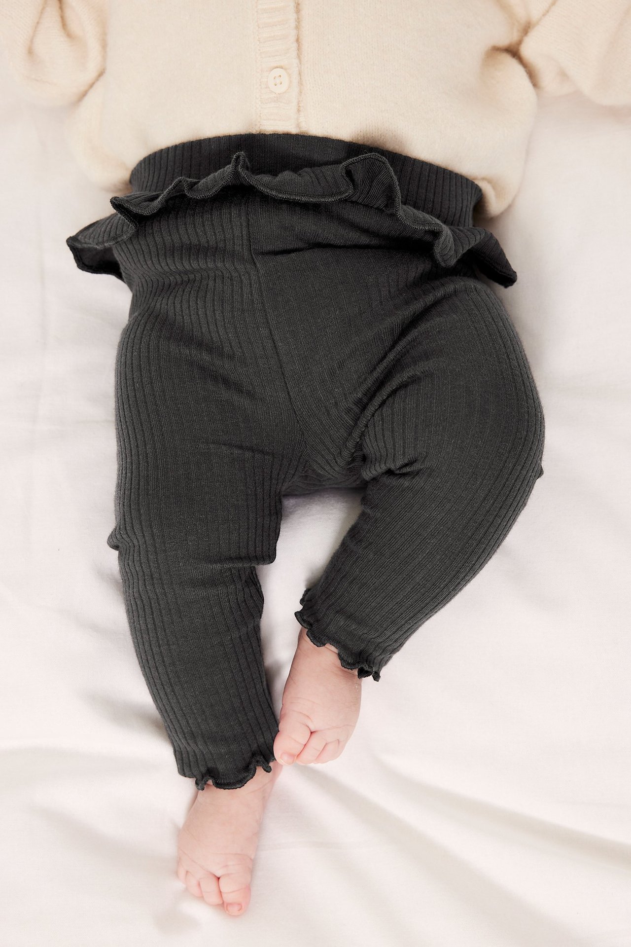 Uppifrån-vy av baby i svarta ribbade leggings med volang i midjan och en ljusbeige kofta.