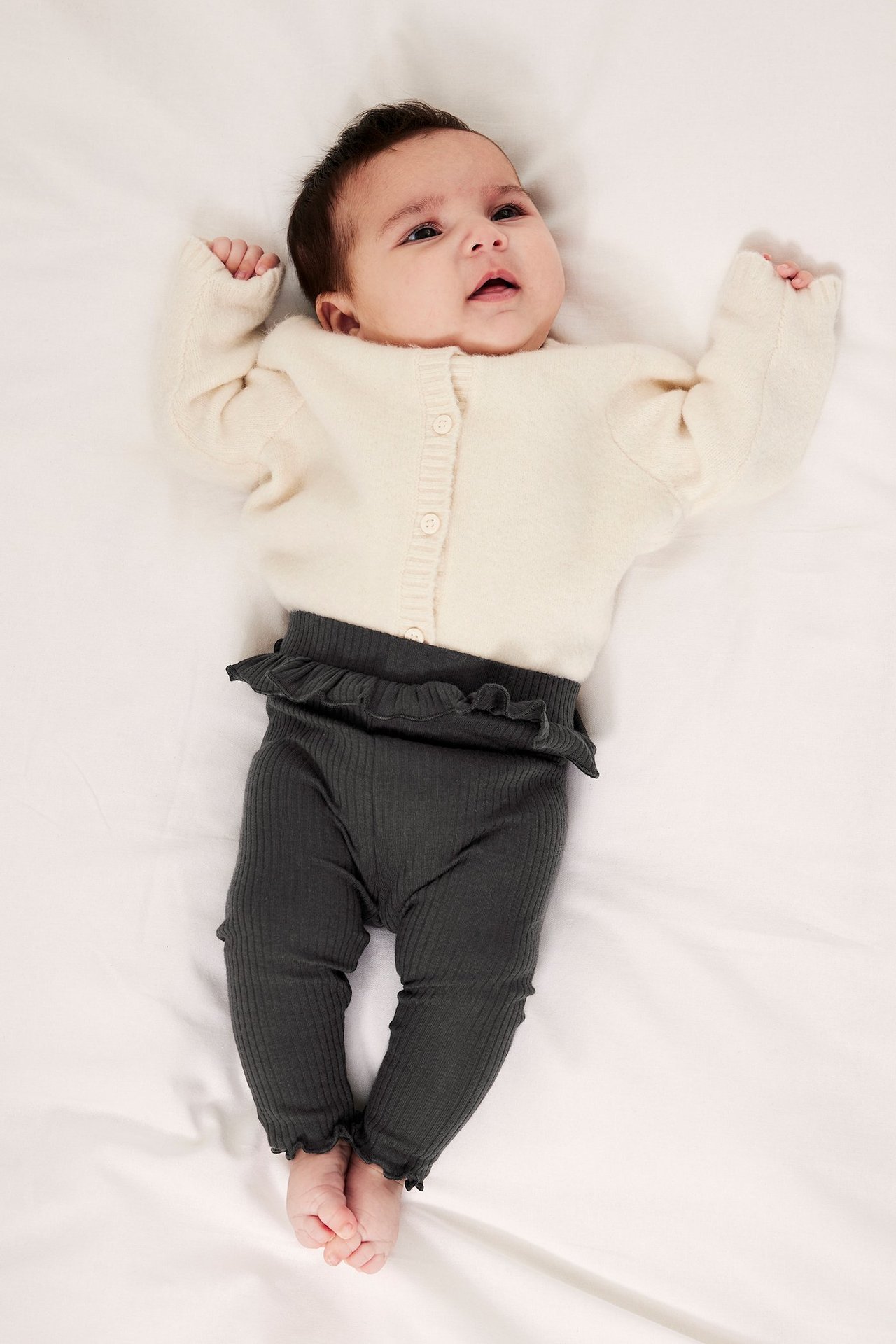 Ovanifrån: baby i mörkgrå ribbade leggings med volang i midjan och en ljusbeige kofta med knappar.