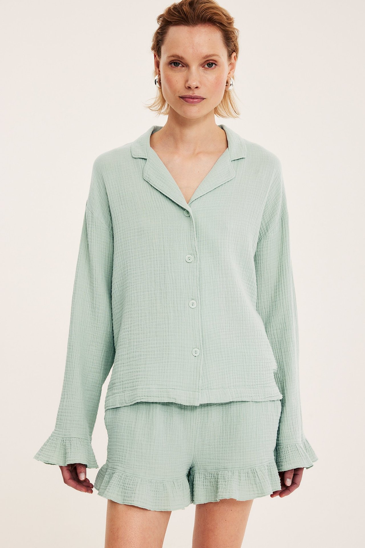 Edestä: Nainen vaaleanvihreässä kuvioidussa puuvillapyjamapaidassa, jossa röyhelöt, ja yhteensopivissa shortseissa.