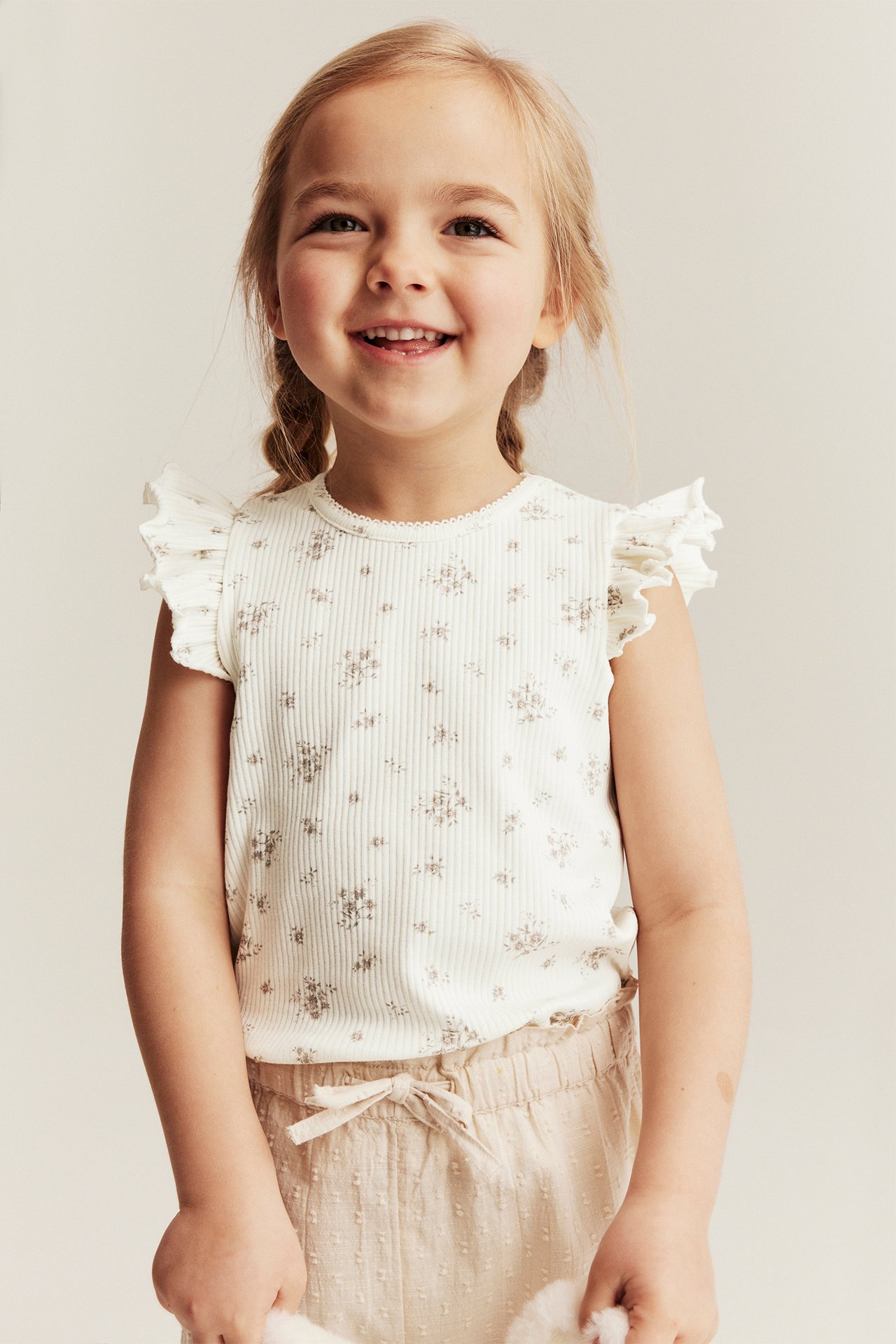 Framifrån: leende barn i offwhite ribbad blommig topp med volangärmar och ljusbeige shorts.