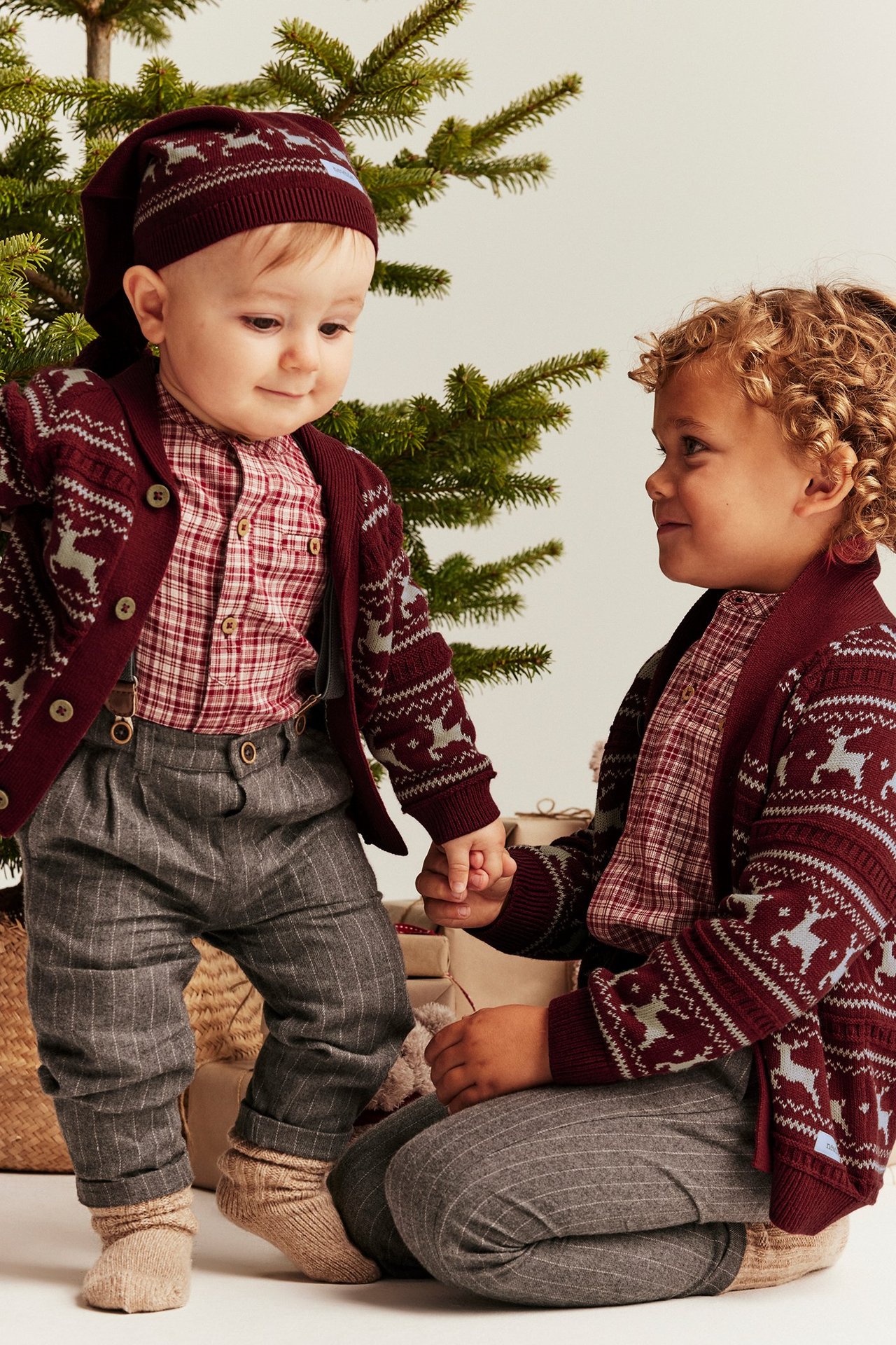 Etunäkymä: Kaksi lasta punaisissa jacquard-neuletakkeissa, ruutupaidat, harmaat housut. Vauvalla yhteensopiva myssy.