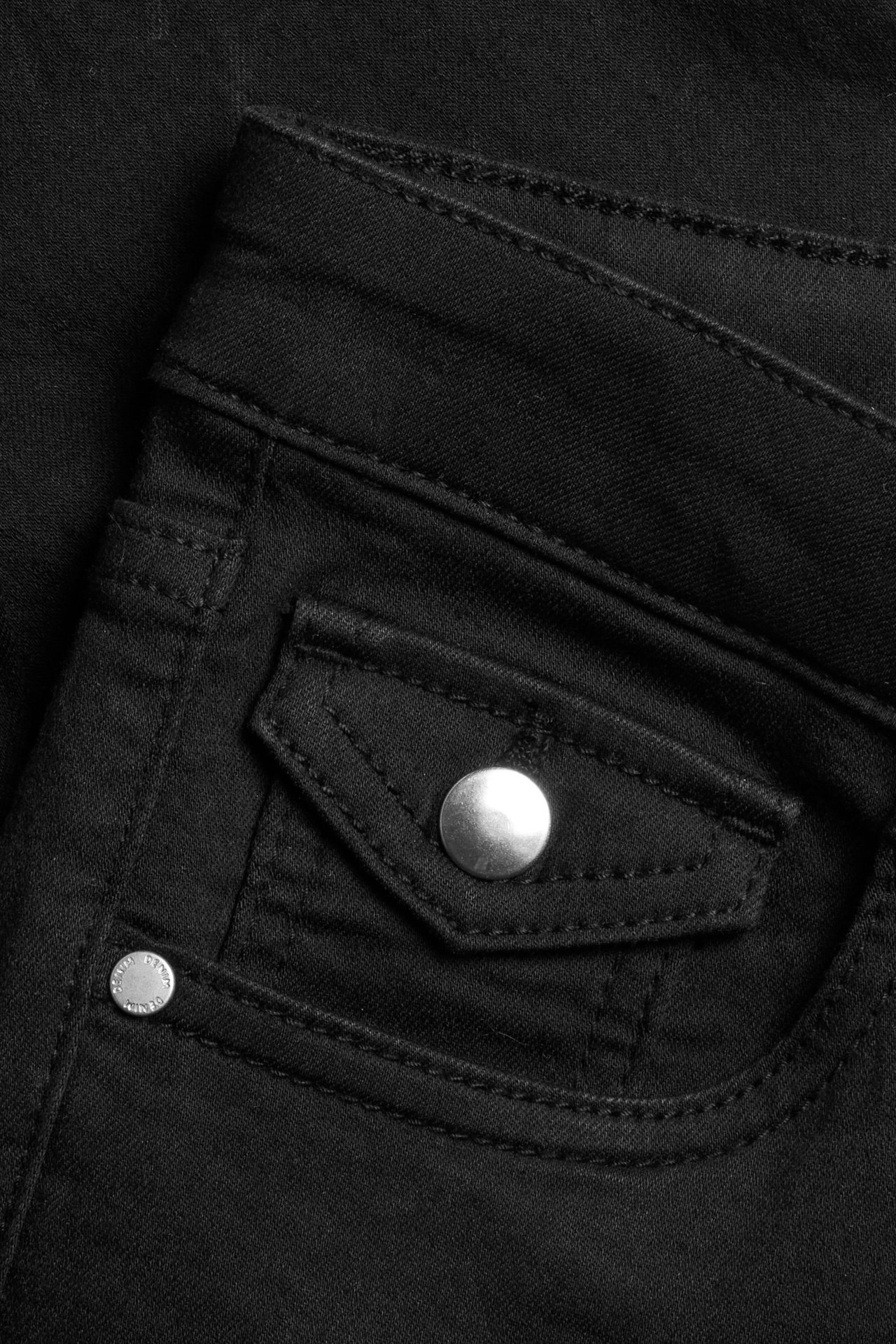 Zbliżenie czarnych jeansów bootcut, widać klapkę kieszeni z srebrnym zatrzaskiem i nity.
