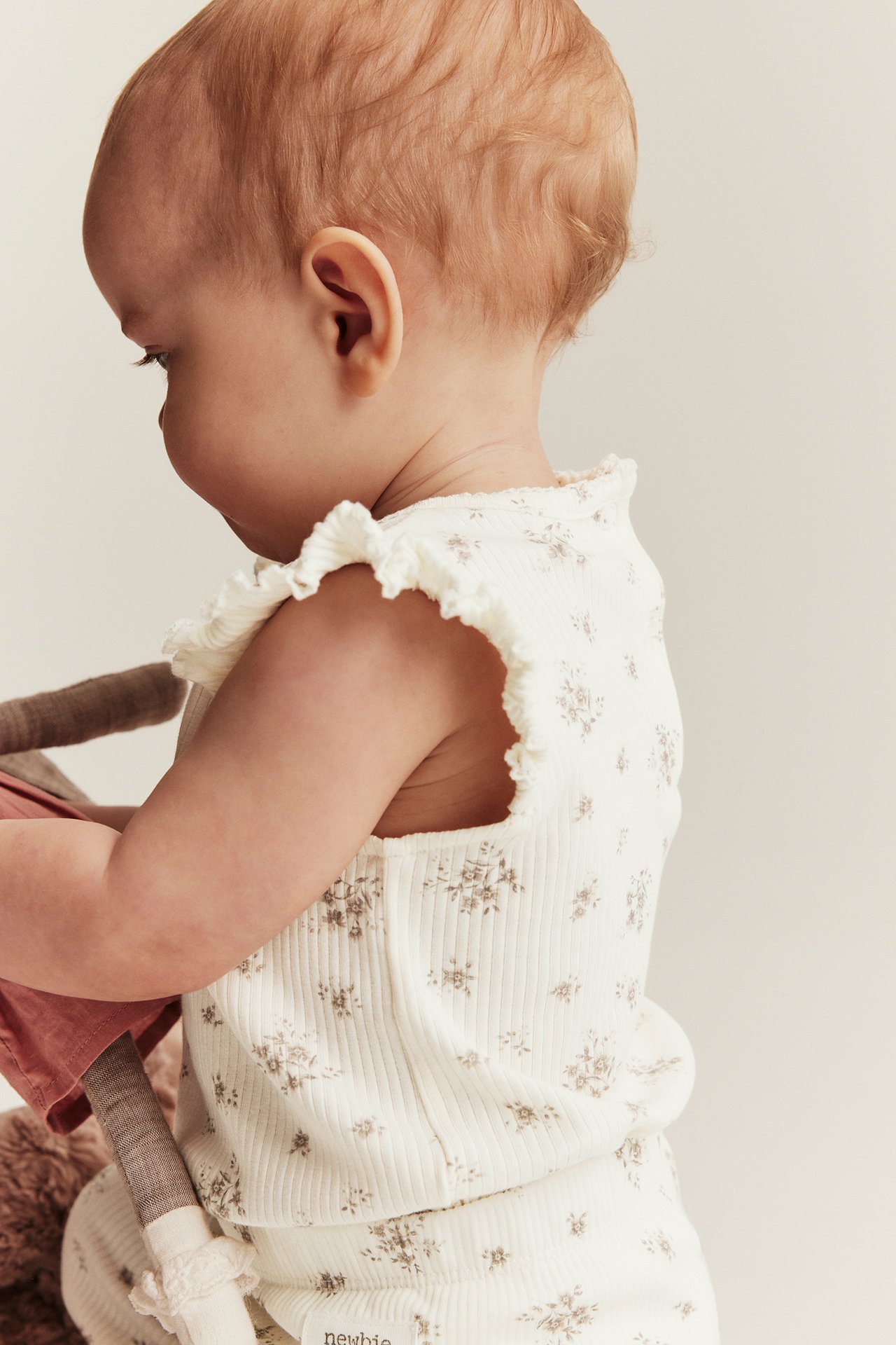 Sidovy av baby i offwhite ribbad blommig body med volangärm, matchande leggings, håller en docka.