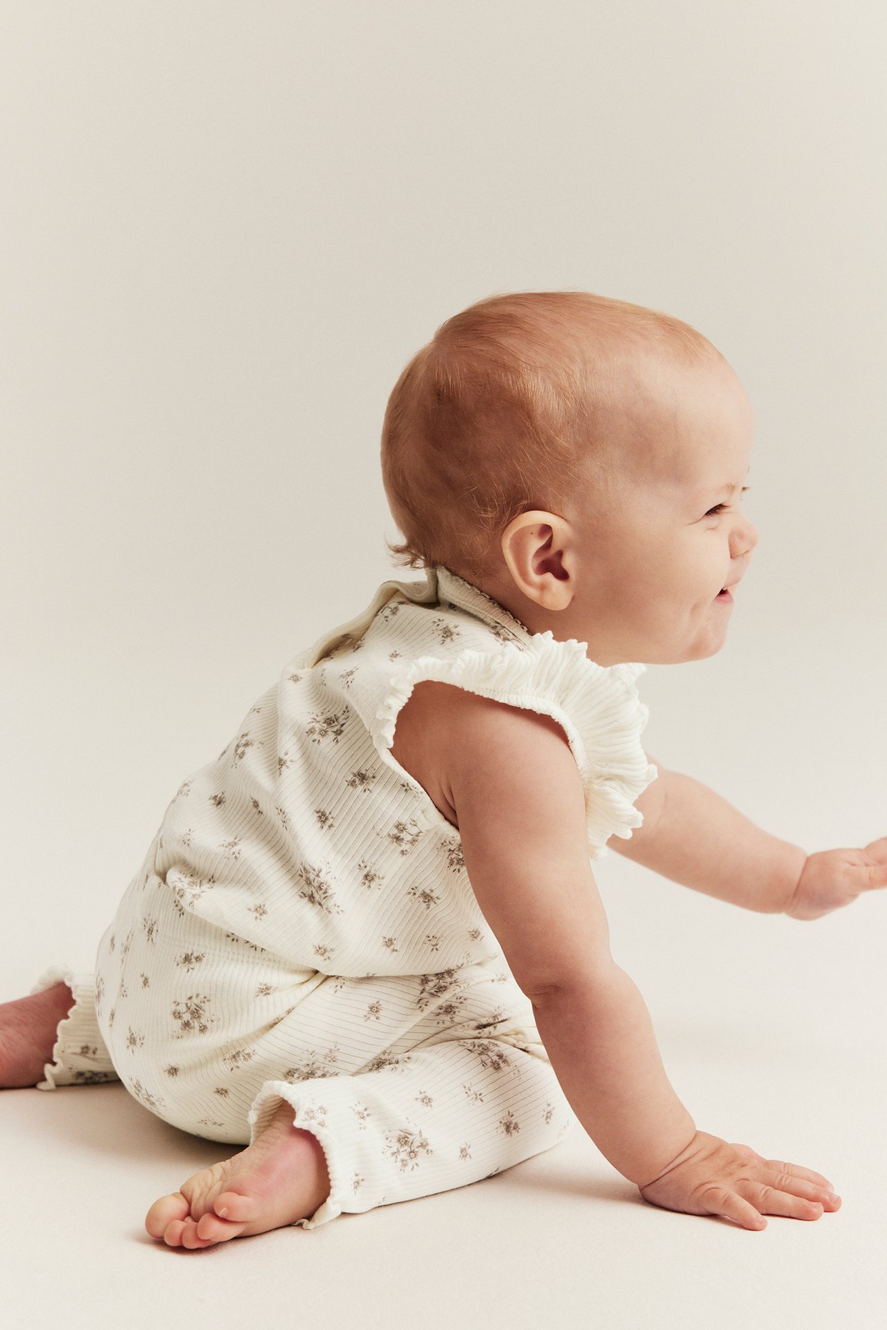 Sidovy av baby i offwhite ribbad blommig body med volangärm och matchande leggings.