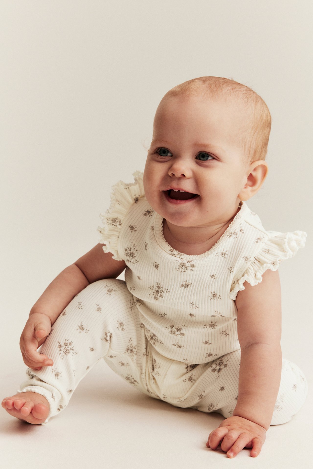 Edestä kuvattu hymyilevä vauva yllään luonnonvalkoinen ribattu kukkakuvioinen body röyhelöhihoilla ja yhteensopivat leggingsit.