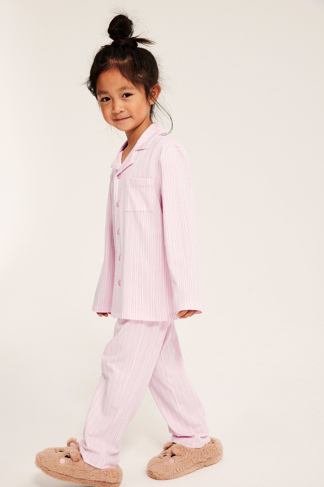 Etukuva lapsesta, yllään pinkki-valkoraidallinen jersey-pyjama napitetulla yläosalla ja ruskeat nalletohvelit.