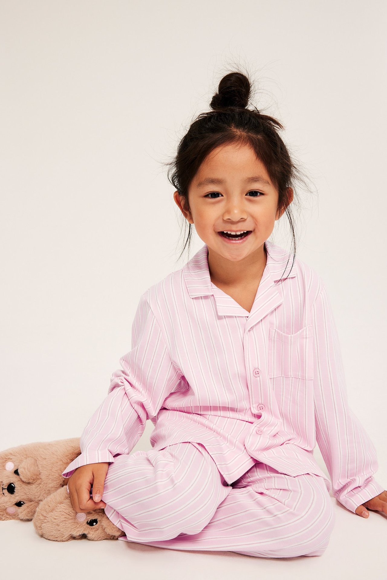 Z przodu, dziecko w różowej piżamie w paski z dżerseju. Góra z kołnierzem, długie spodnie, brązowe kapcie.