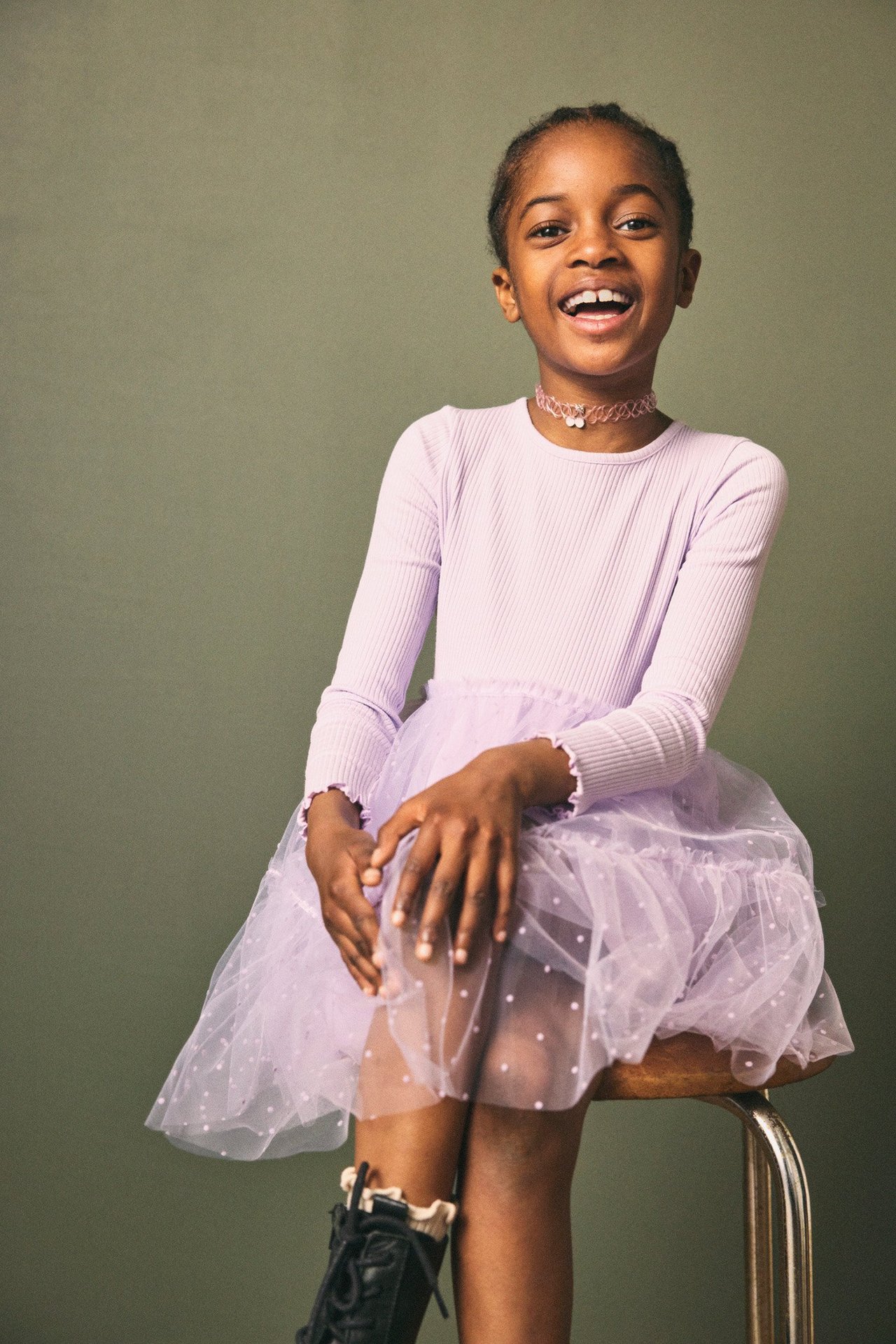 Z przodu: Dziecko w jasnofioletowej sukience z prążkowaną górą i tiulową spódnicą, różowym chokerem, czarnymi butami.