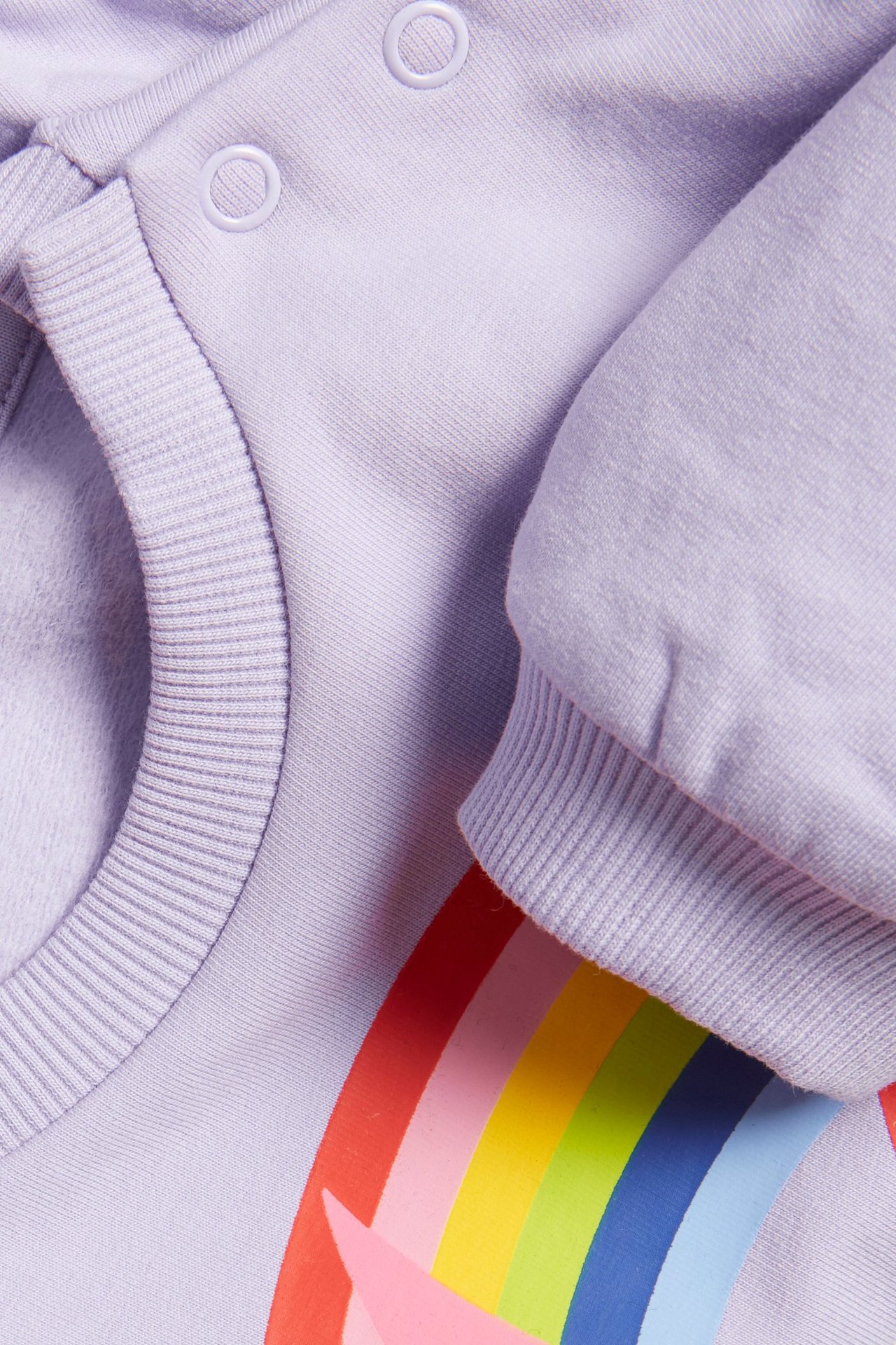 Zbliżenie na jasnofioletową bluzę niemowlęcą z kolorowym nadrukiem Babblarna i zatrzaskami na ramieniu.