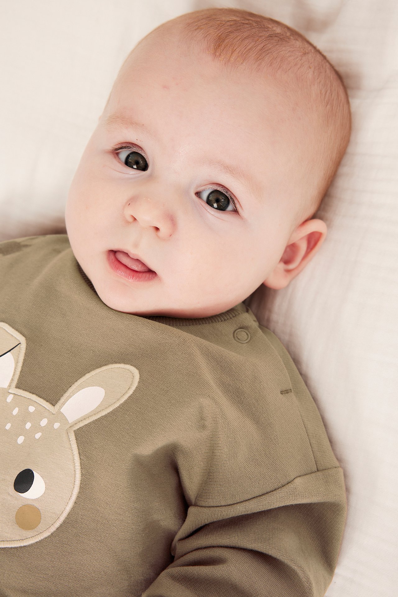 Närbild på en baby i en brun bomullssweatshirt med rådjursapplikation och tryckknappar på axeln.