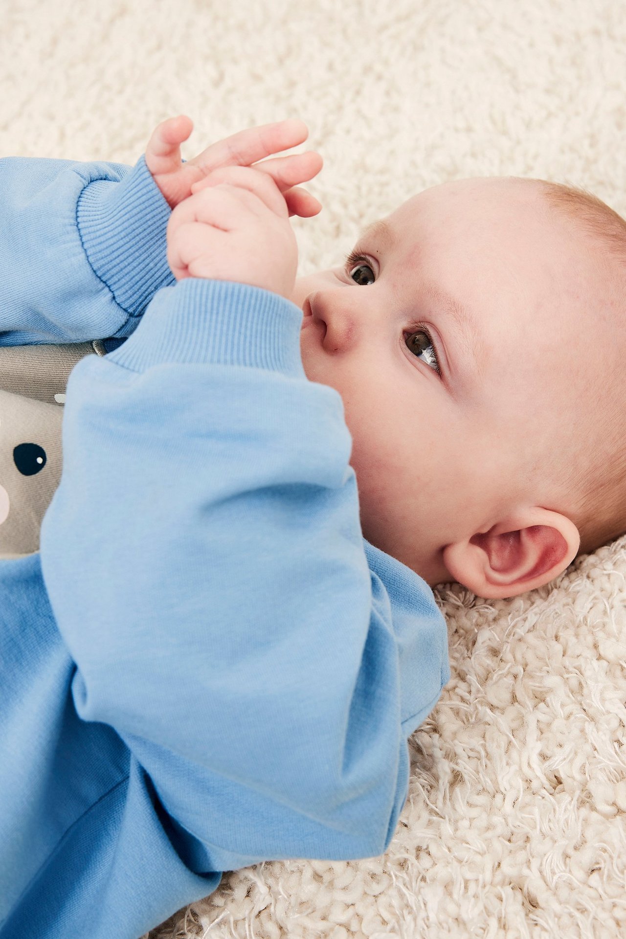Zbliżenie na dziecko w niebieskiej bluzie z aplikacją zwierzęcą, leżące na białym dywanie.