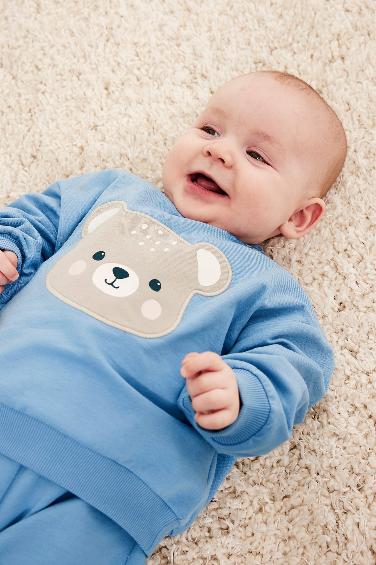 Widok z przodu uśmiechniętego dziecka w niebieskiej bawełnianej bluzie z aplikacją misia i zatrzaskami.