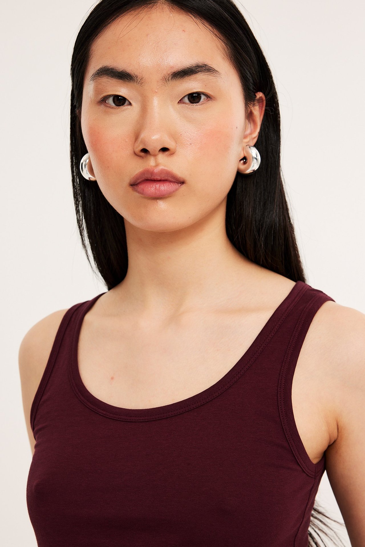 Nærbilde forfra av en kvinne i mørkerød bomullsblanding singlet og sølvfargede øreringer.