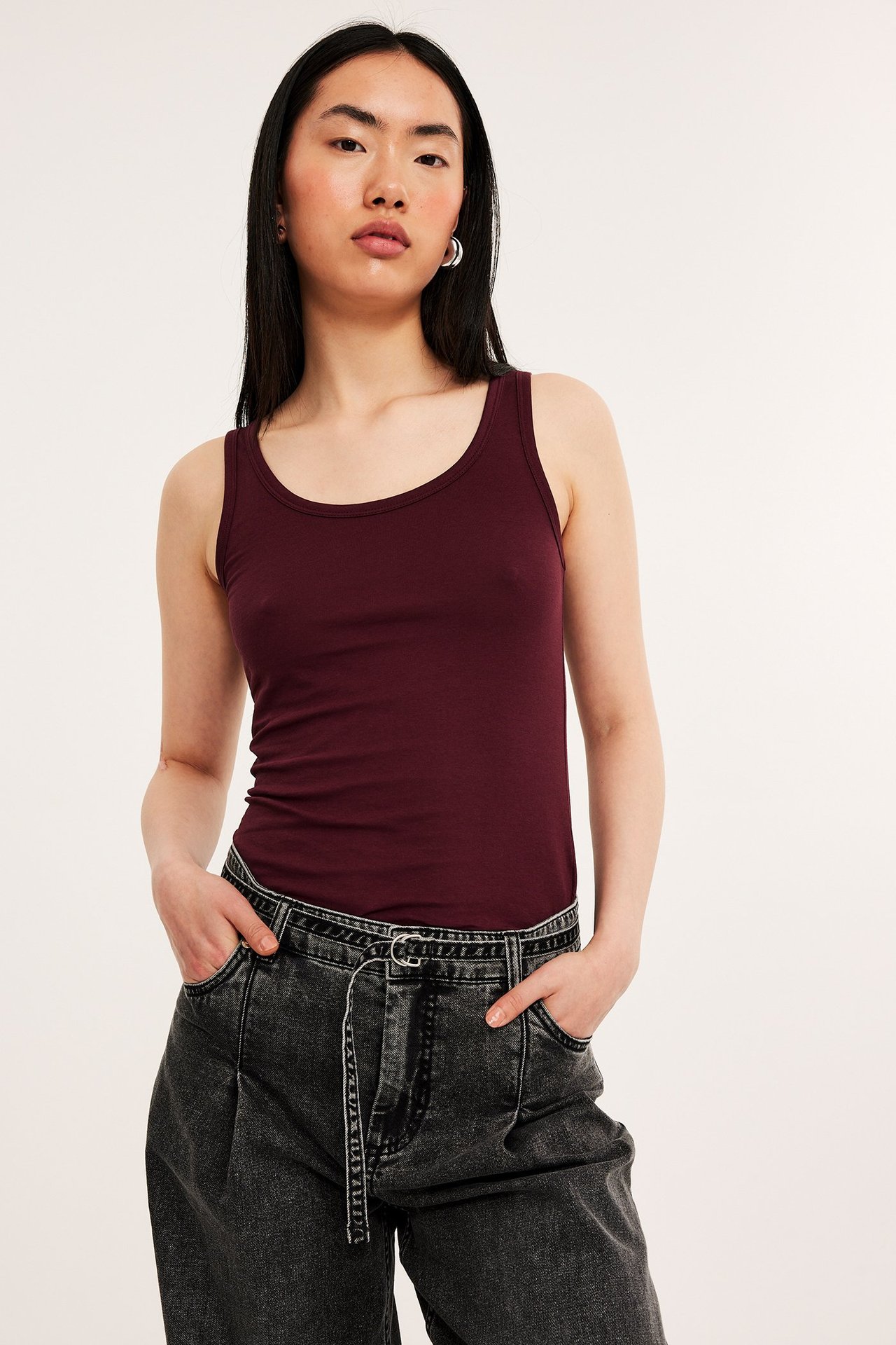 Forfra: kvinne i mørkerød figurnær singlet og mørkegrå jeans med hendene i lommene.