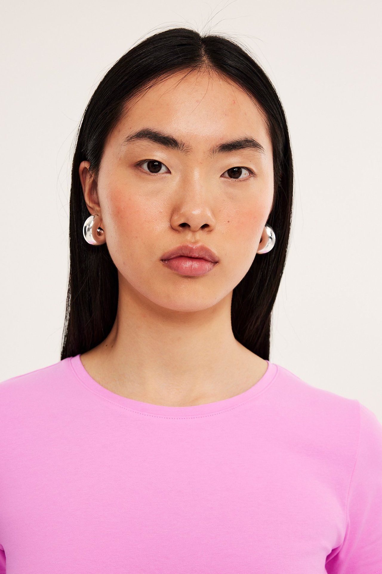 Nærbilde forfra av kvinne i rosa kortermet rundhalset basetopp og sølvfargede kuppelformede øredobber.