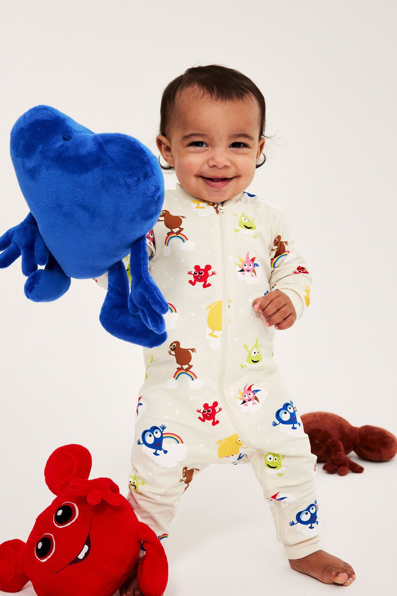 Leende baby i ljusbeige mönstrad Babblarna-pyjamas med dragkedja fram, håller en blå mjukisleksak.
