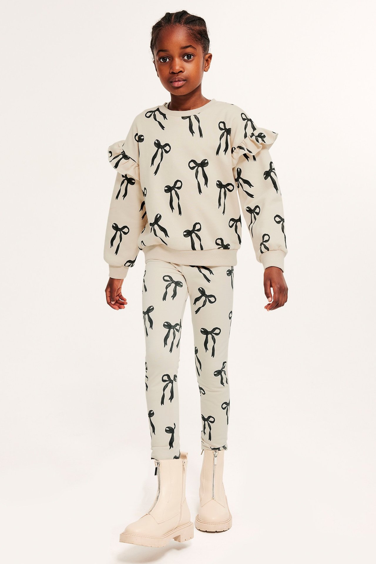 Dziecko w jasnobeżowej bluzie z wzorem kokardek i falbankami na rękawach, pasujących legginsach i beżowych botkach.