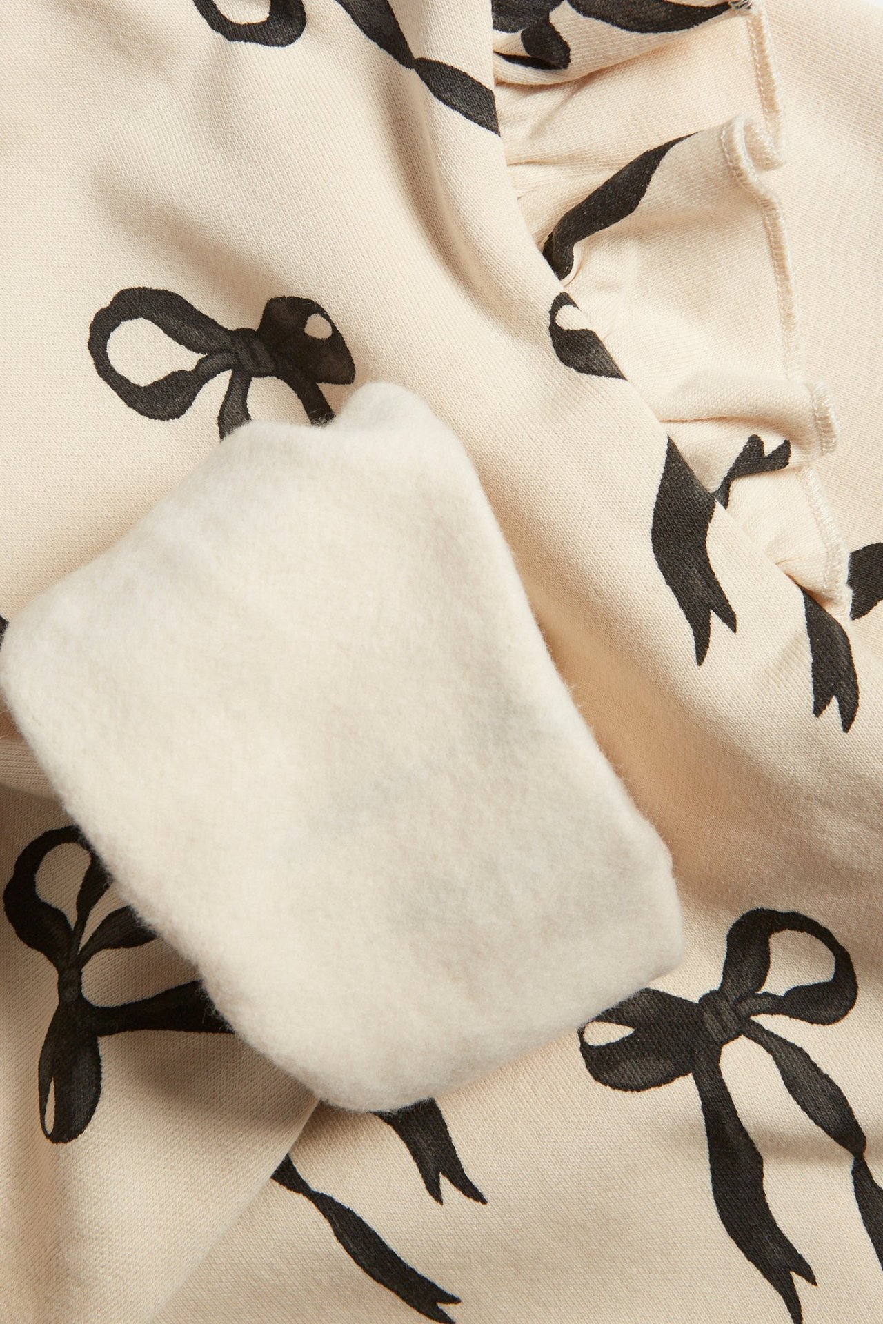 Zbliżenie na jasnobeżową bluzę dziecięcą z czarnym wzorem kokardek i falbankami na rękawie.