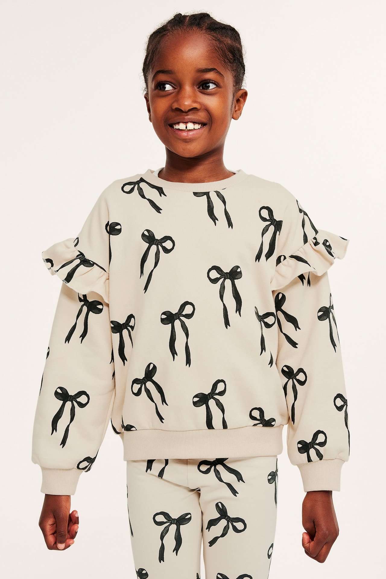 Widok z przodu: Dziecko w jasnobeżowej bluzie z czarnym wzorem kokardek, falbankami na ramionach i pasującymi spodniami.