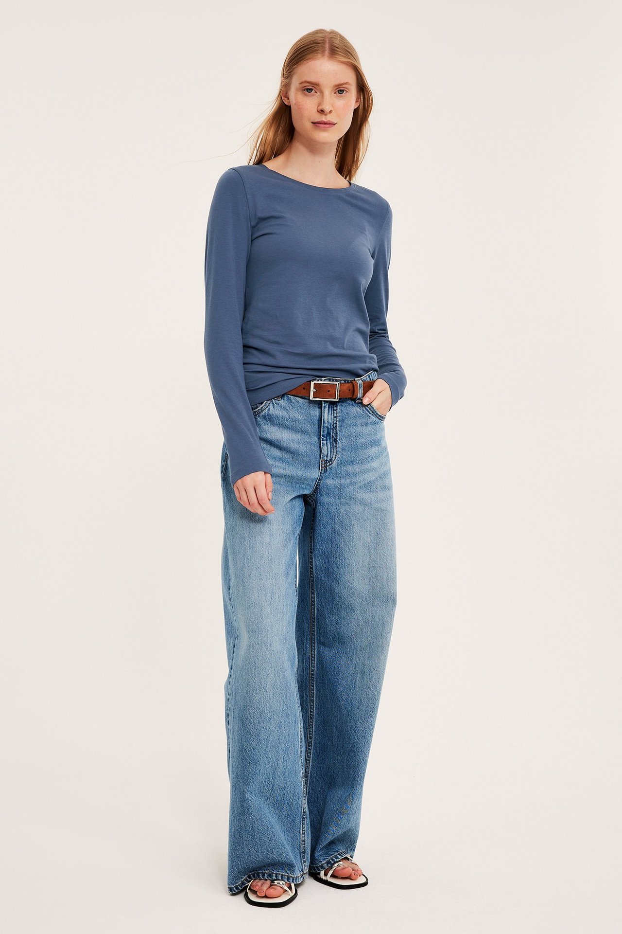 Forfra: Kvinne i blå langermet basetopp, lyseblå vide jeans, brunt belte og hvite sandaler.