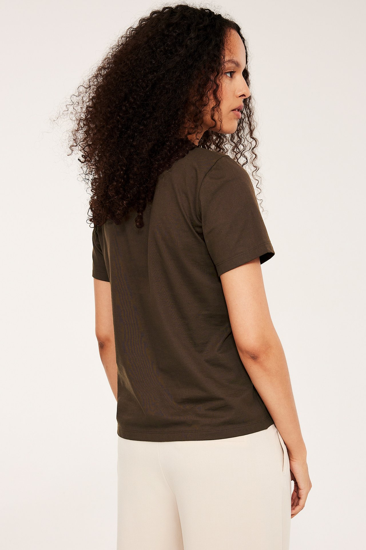 Bakfra høyre: kvinne i en mørk brun, ensfarget bomulls-t-skjorte med rett passform og lyse beige bukser.