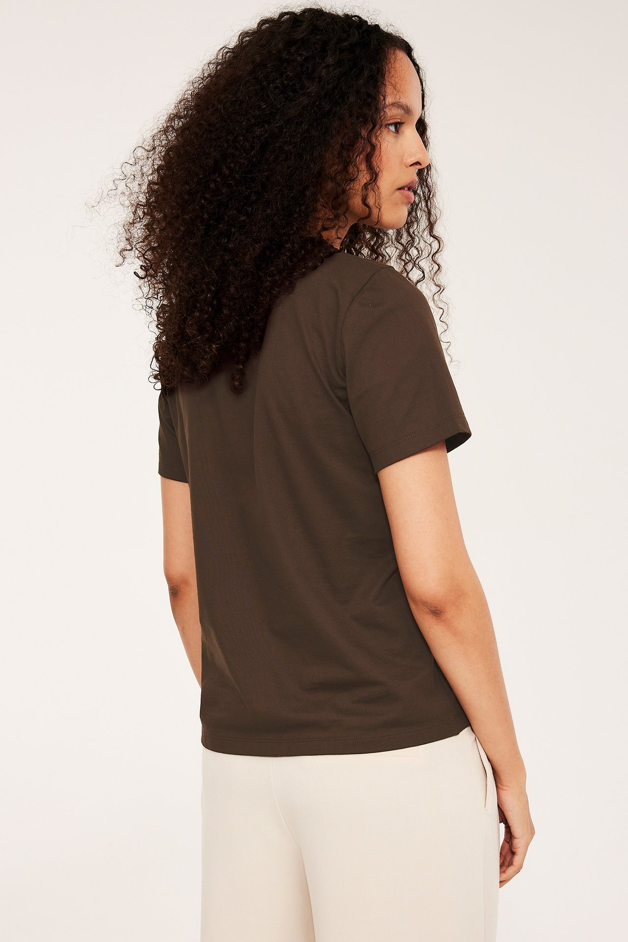 Widok z boku/tyłu kobiety w ciemnobrązowej, prostej bawełnianej koszulce i jasnobeżowych spodniach.