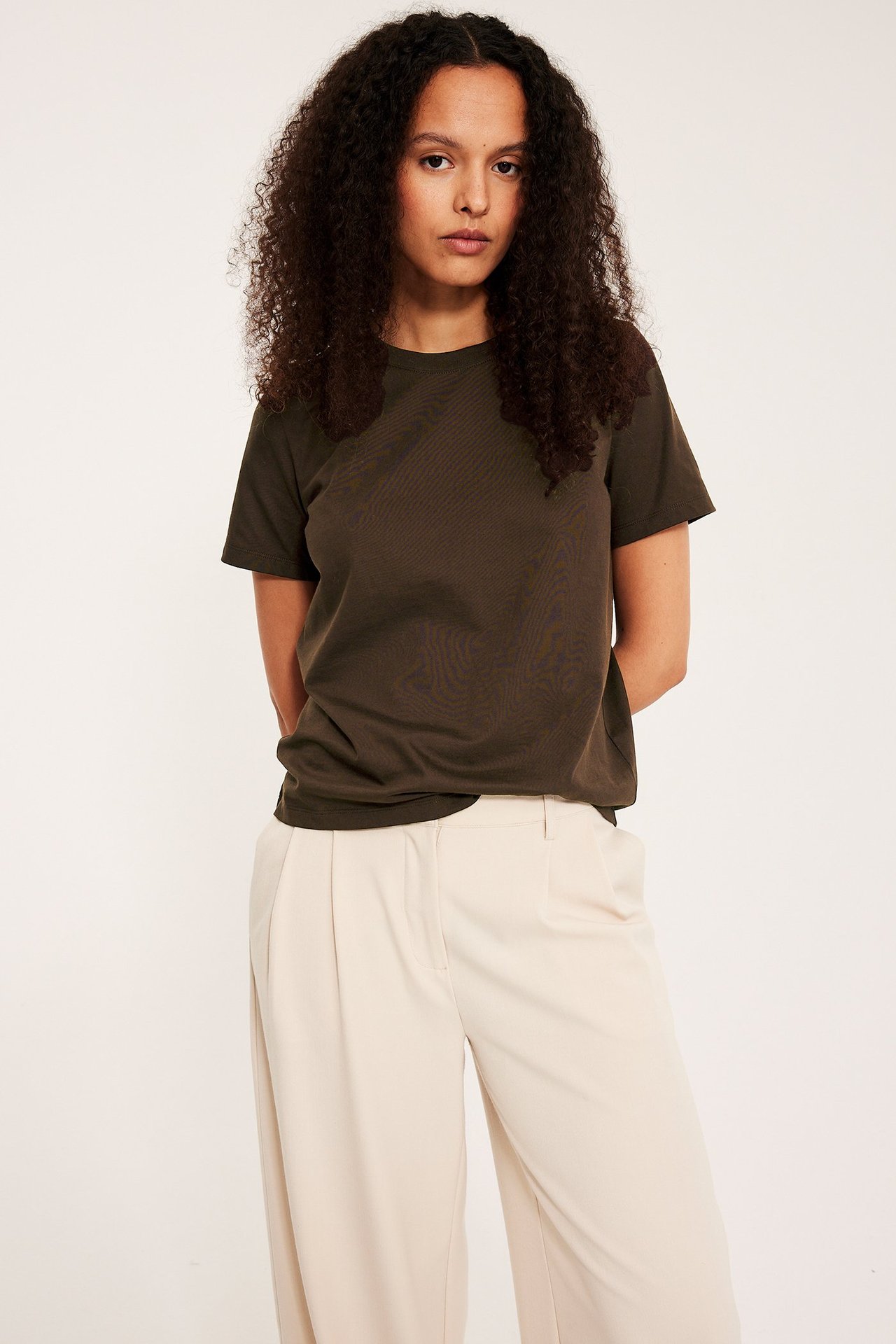 Forfra, kvinne i mørkebrun bomulls-T-skjorte og beige plisserte vide bukser.