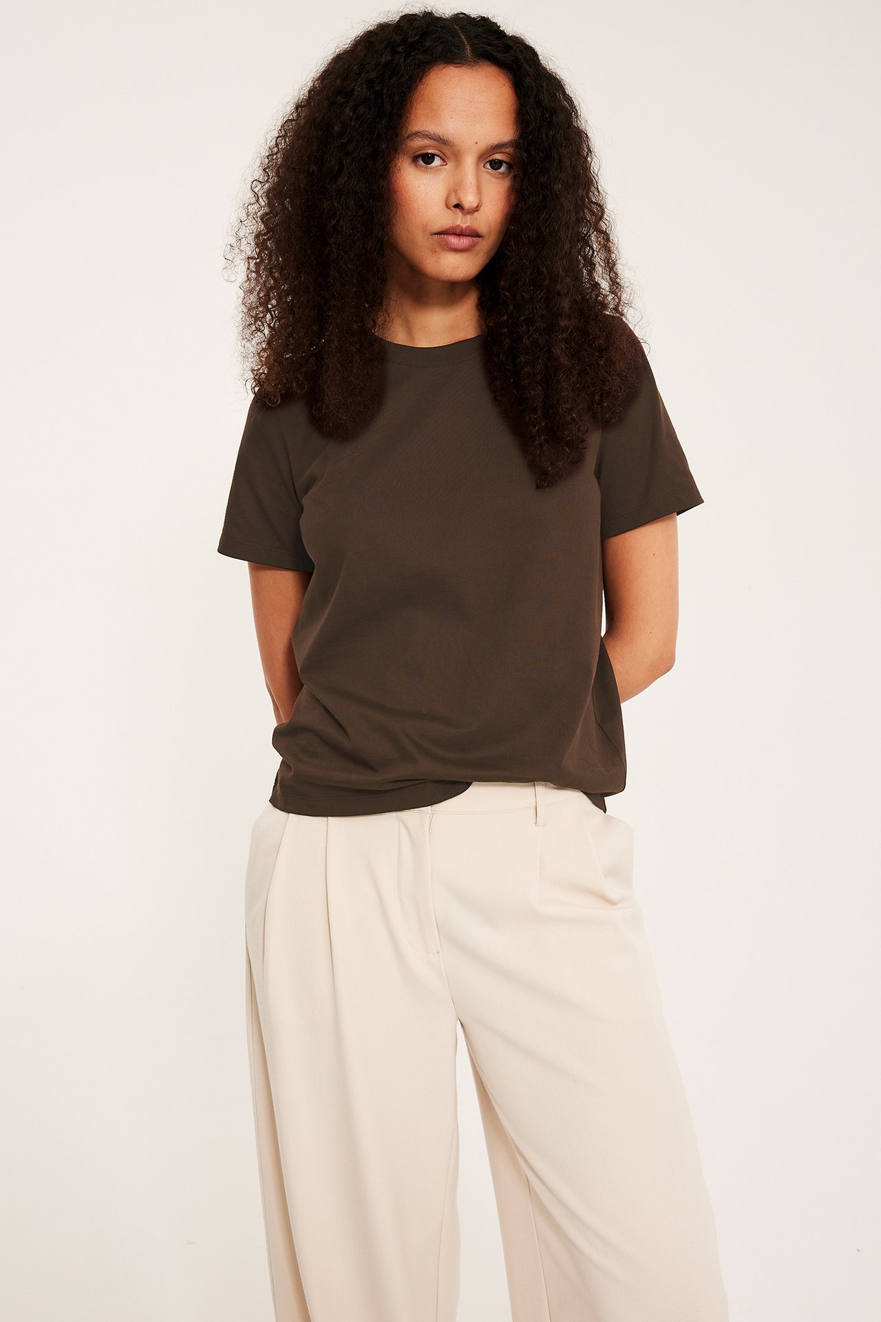 Widok z przodu: kobieta w ciemnobrązowym bawełnianym T-shircie i jasnobeżowych szerokich spodniach.