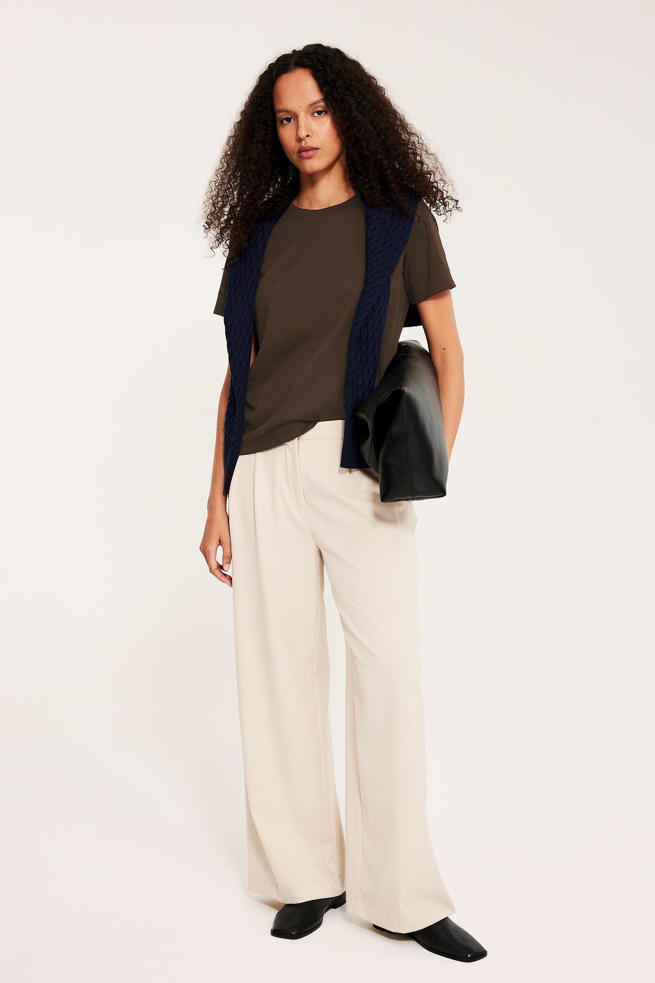 Widok z przodu: Kobieta w ciemnobrązowym T-shircie, beżowych spodniach, granatowym swetrze, czarnych butach, trzyma czarną torbę.