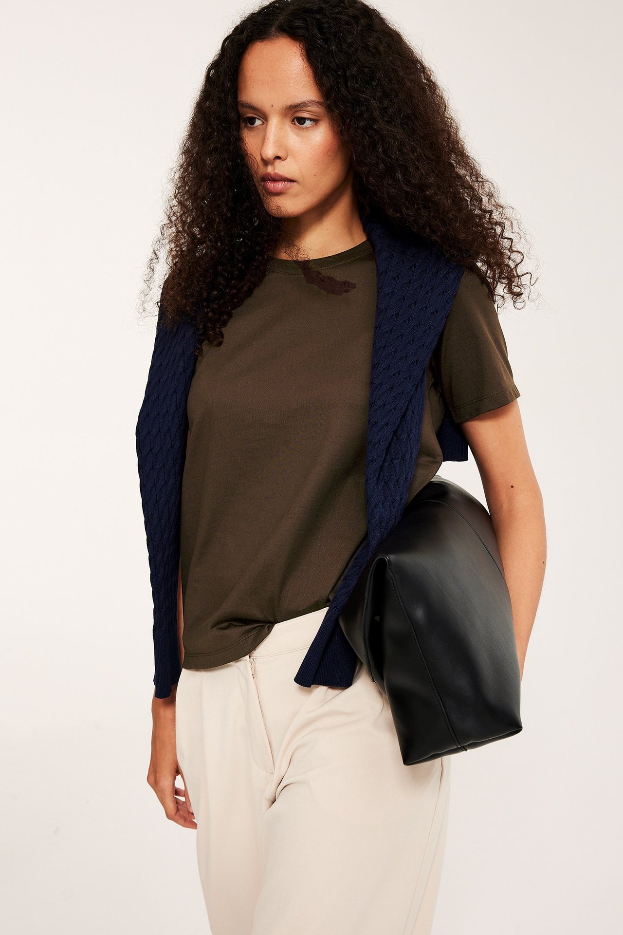 Frontvisning: Kvinne i en mørkebrun bomulls-T-skjorte, lysebeige bukser, marineblå genser og svart veske.