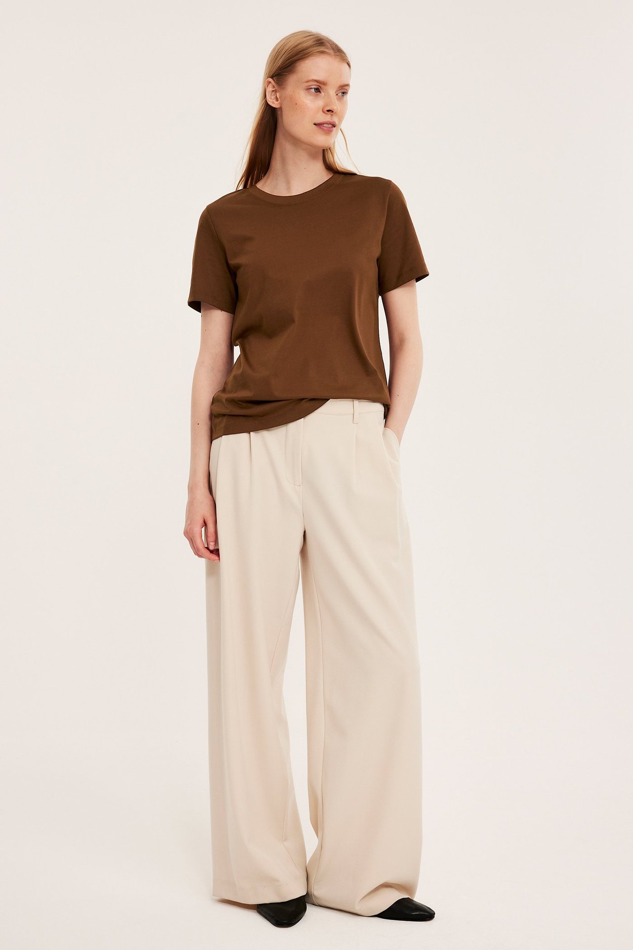 Forfra: Kvinne i brun bomulls-T-skjorte, lysebeige vide bukser og svarte flate sko.