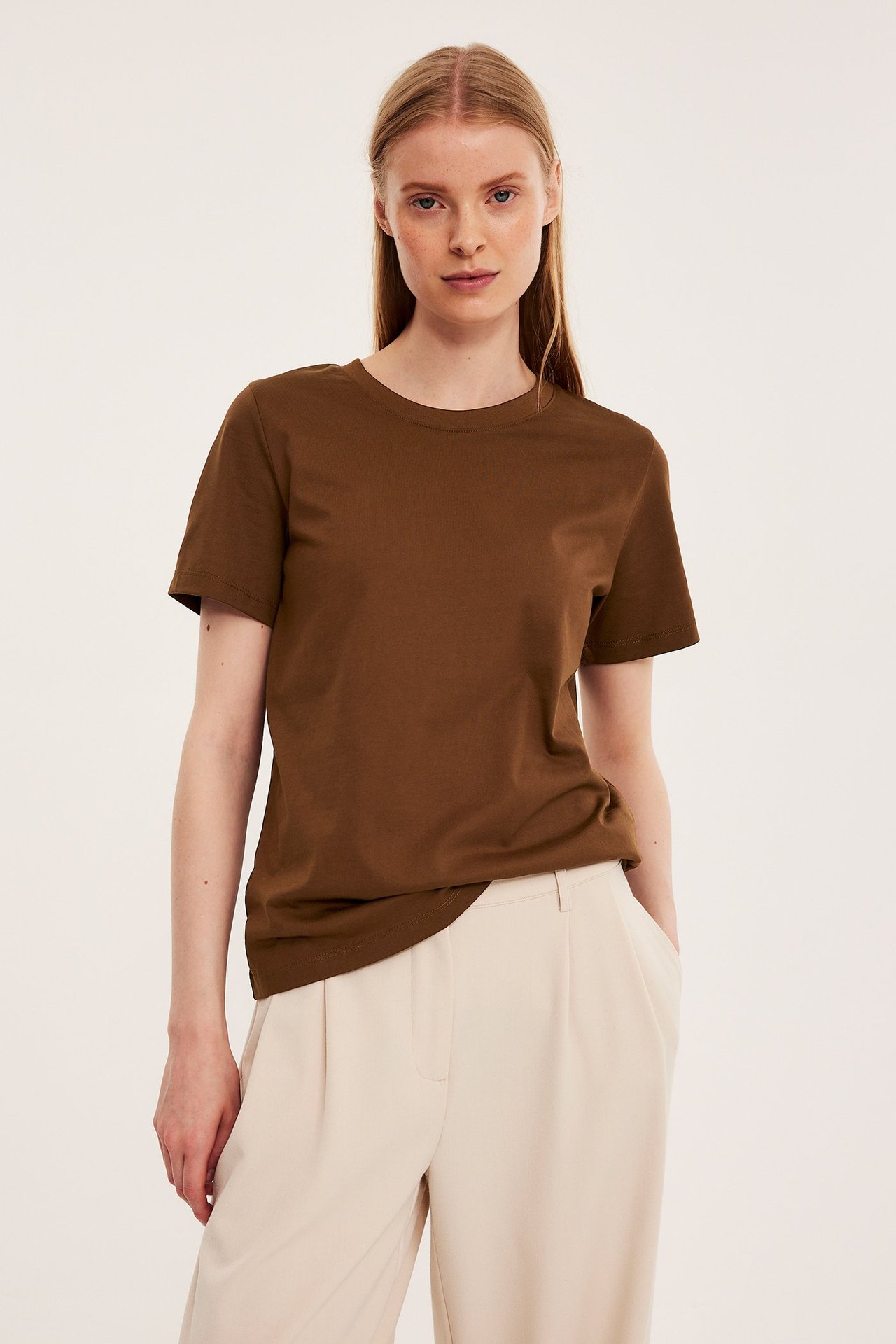 Frontvisning av en kvinne i en brun, rett passform bomulls-T-skjorte og lysebeige plisserte bukser.