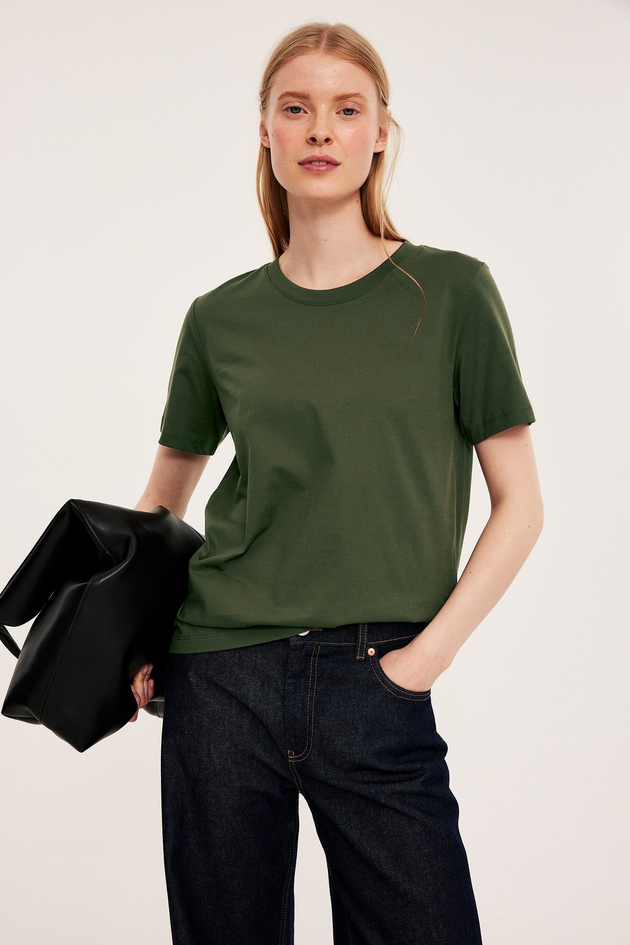 Forfra: kvinne i rett grønn bomulls-T-skjorte, mørkeblå jeans, holder en svart håndveske.