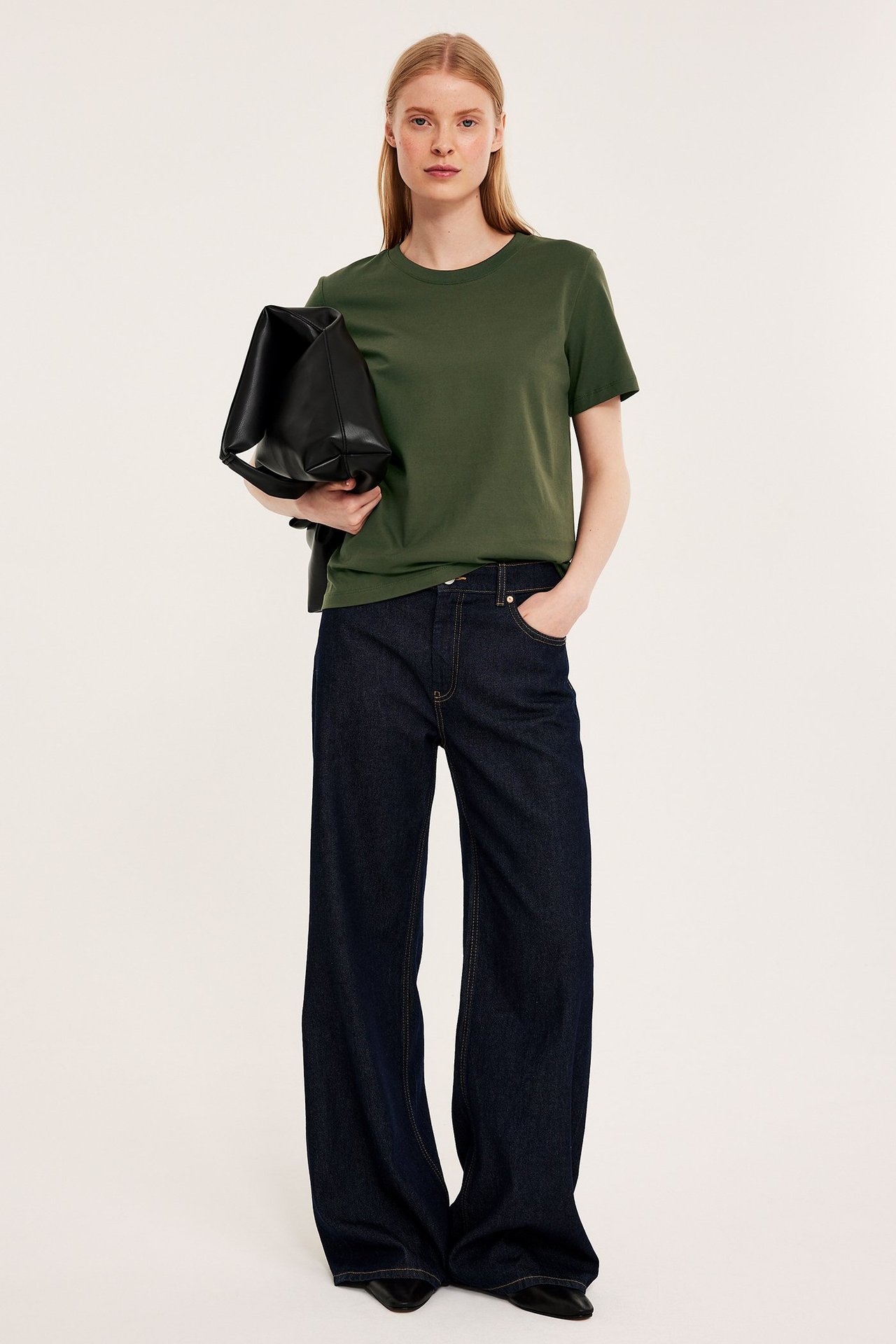 Forfra: kvinne i rett grønn bomulls-T-skjorte, mørkeblå vide jeans og holder en svart håndveske.
