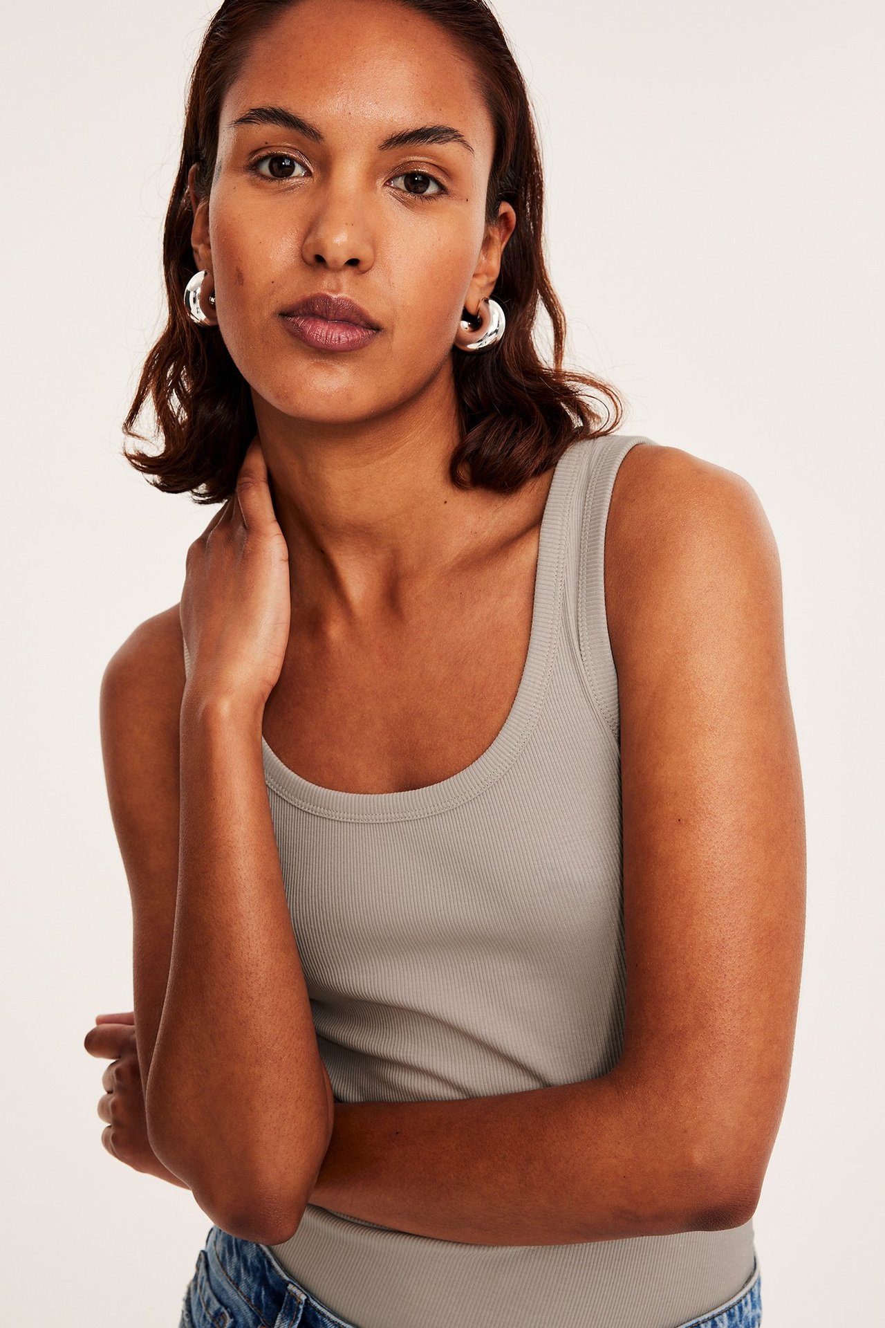 Frontbilde av en kvinne i en figurnær beige ribbet singlet med sølvfargede ringøredobber.
