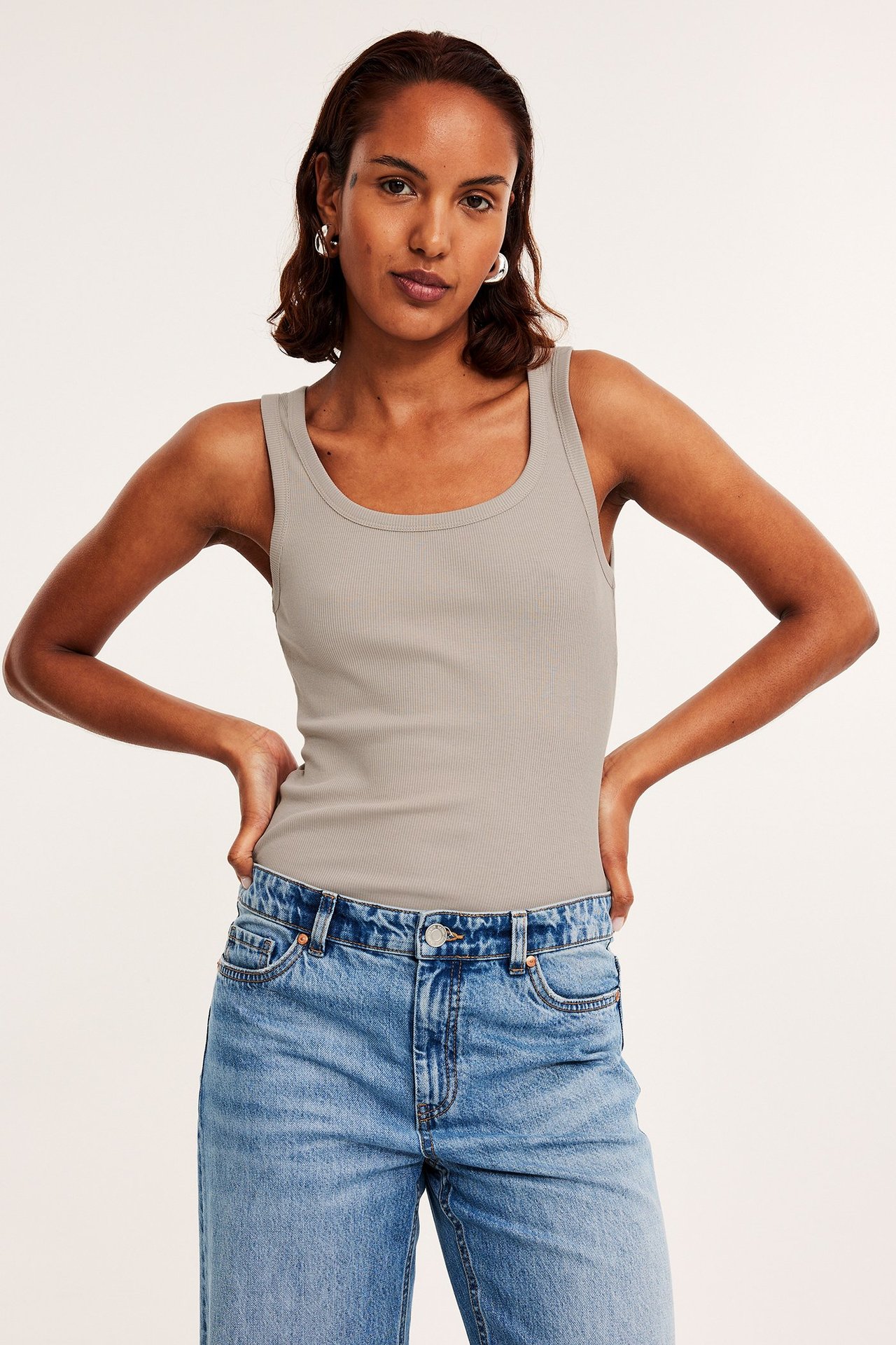 Frontbilde av en kvinne i en figurnær beige ribbet singlet, lyseblå jeans og sølvfargede ringøredobber.