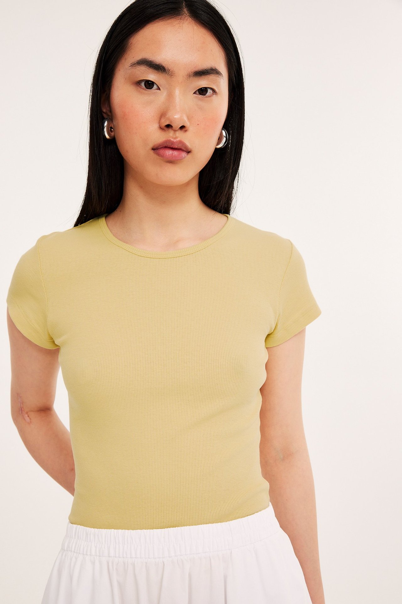 Frontbilde av kvinne i lys gul tettsittende ribbet t-skjorte, hvite bukser og sølvfargede ringøreringer.