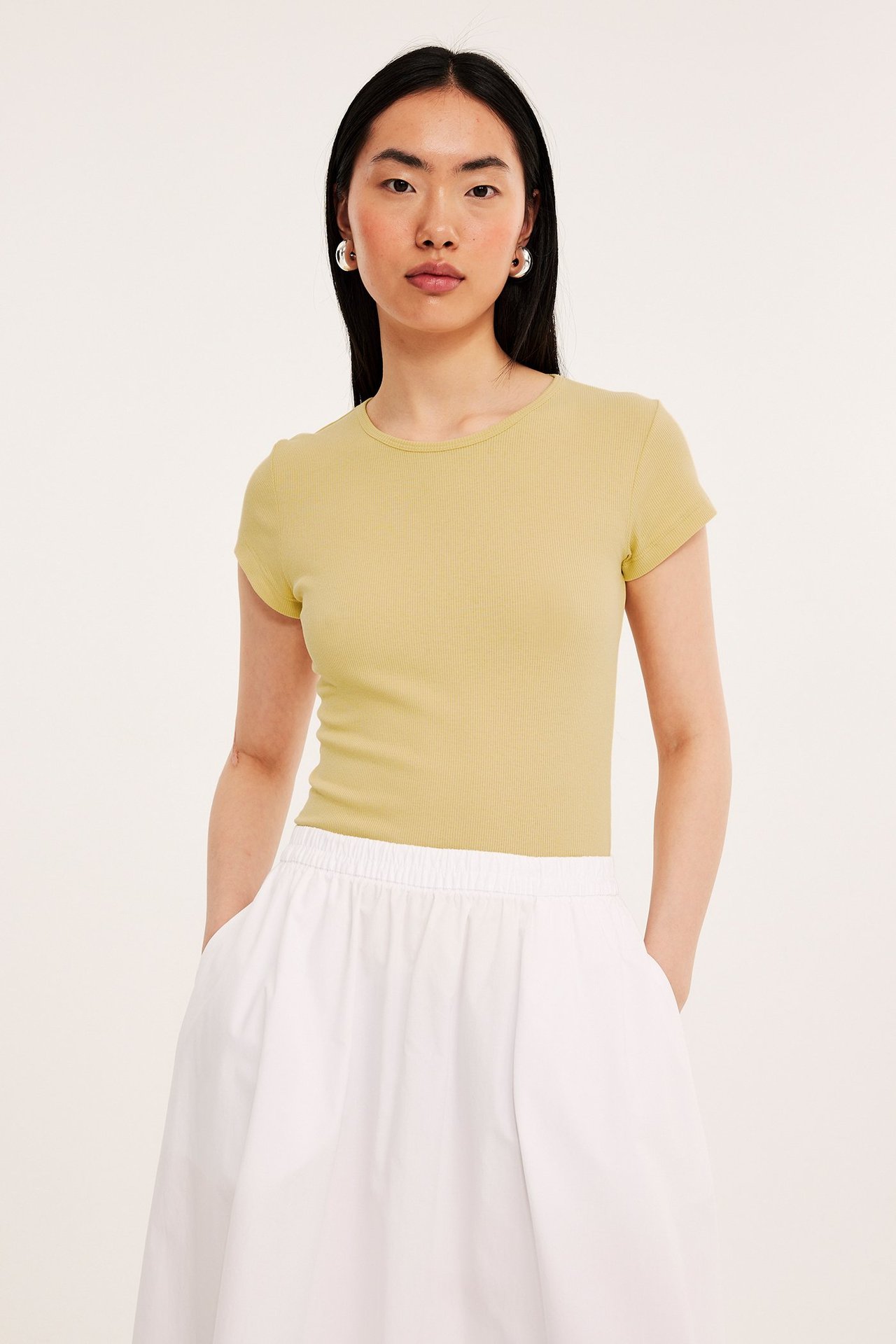 Frontbilde av kvinne i lys gul ribbet kortermet t-skjorte, hvite vide bukser og sølvfargede øreringer.