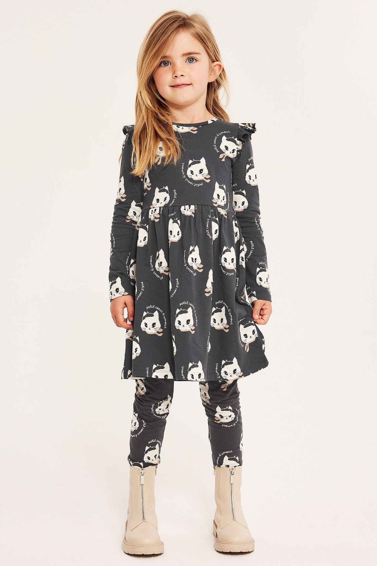 Front view: Kid i mørkegrå bomullsjerseykjole med kattemønster, skulderkapper, matchende leggings og beige støvler.