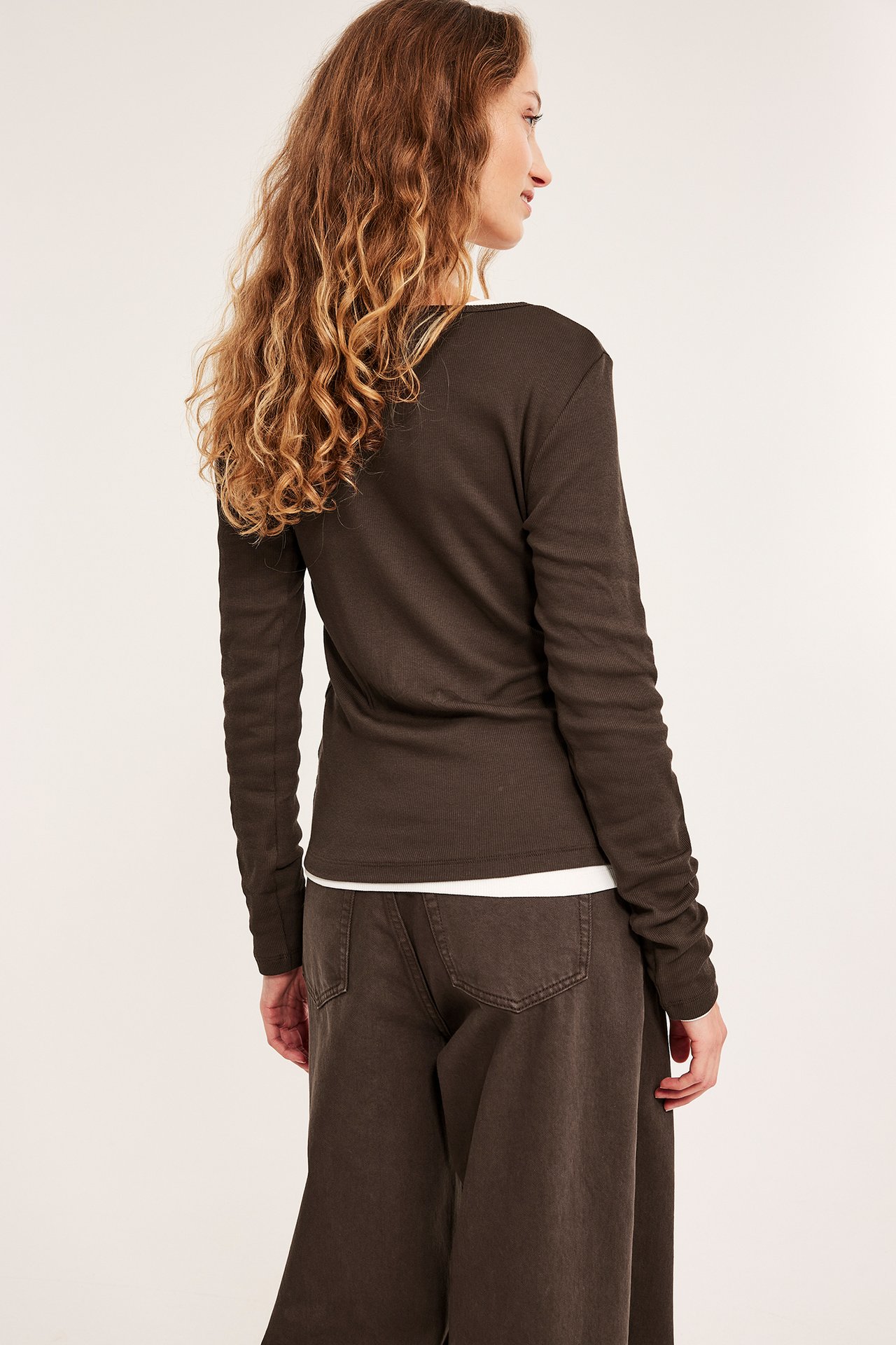Widok z tyłu: Kobieta w ciemnobrązowej prążkowanej bluzce, białym t-shircie i ciemnobrązowych szerokich spodniach.