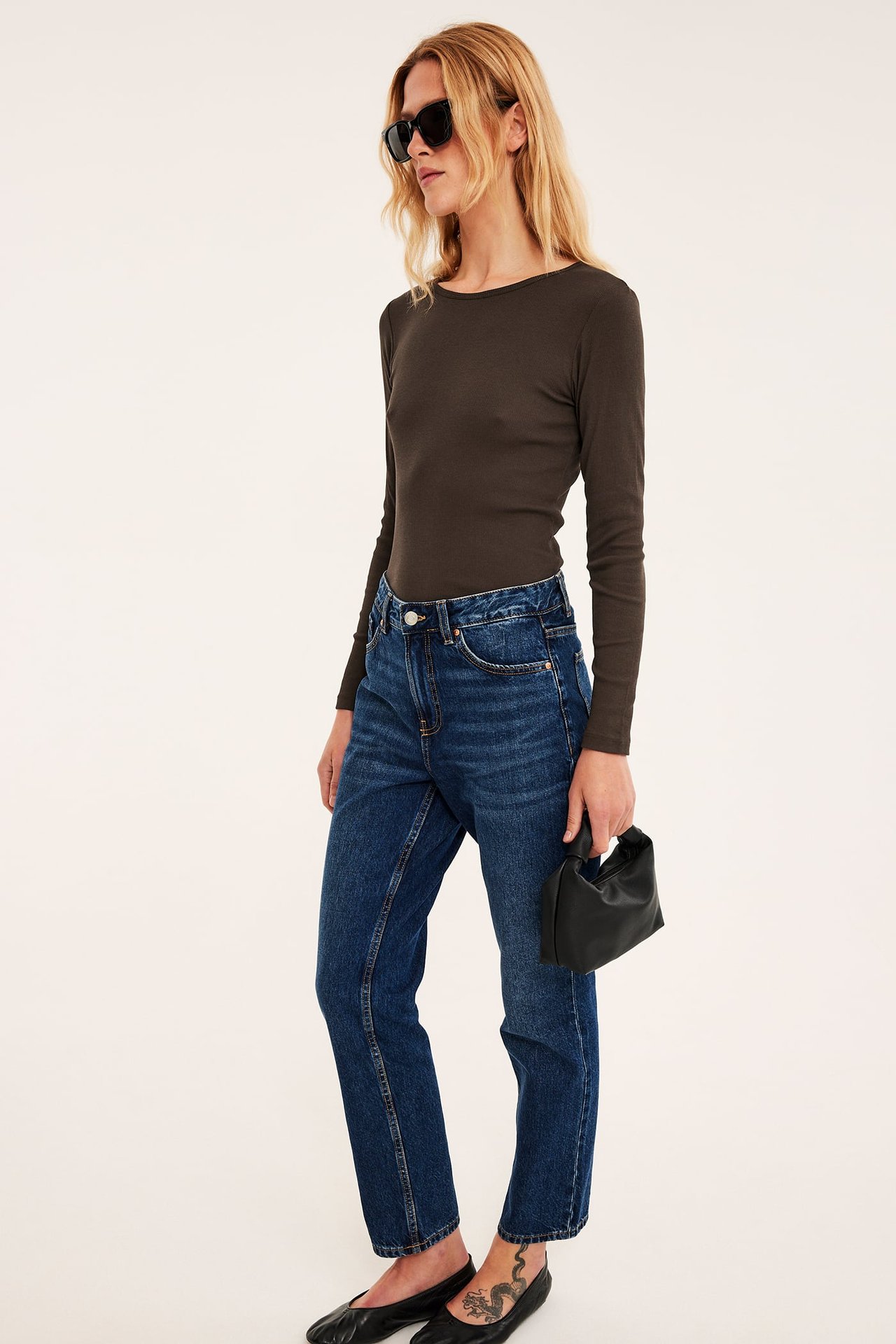 Kvinne i blå rette jeans, mørkebrun langermet topp, svarte solbriller og en svart veske.