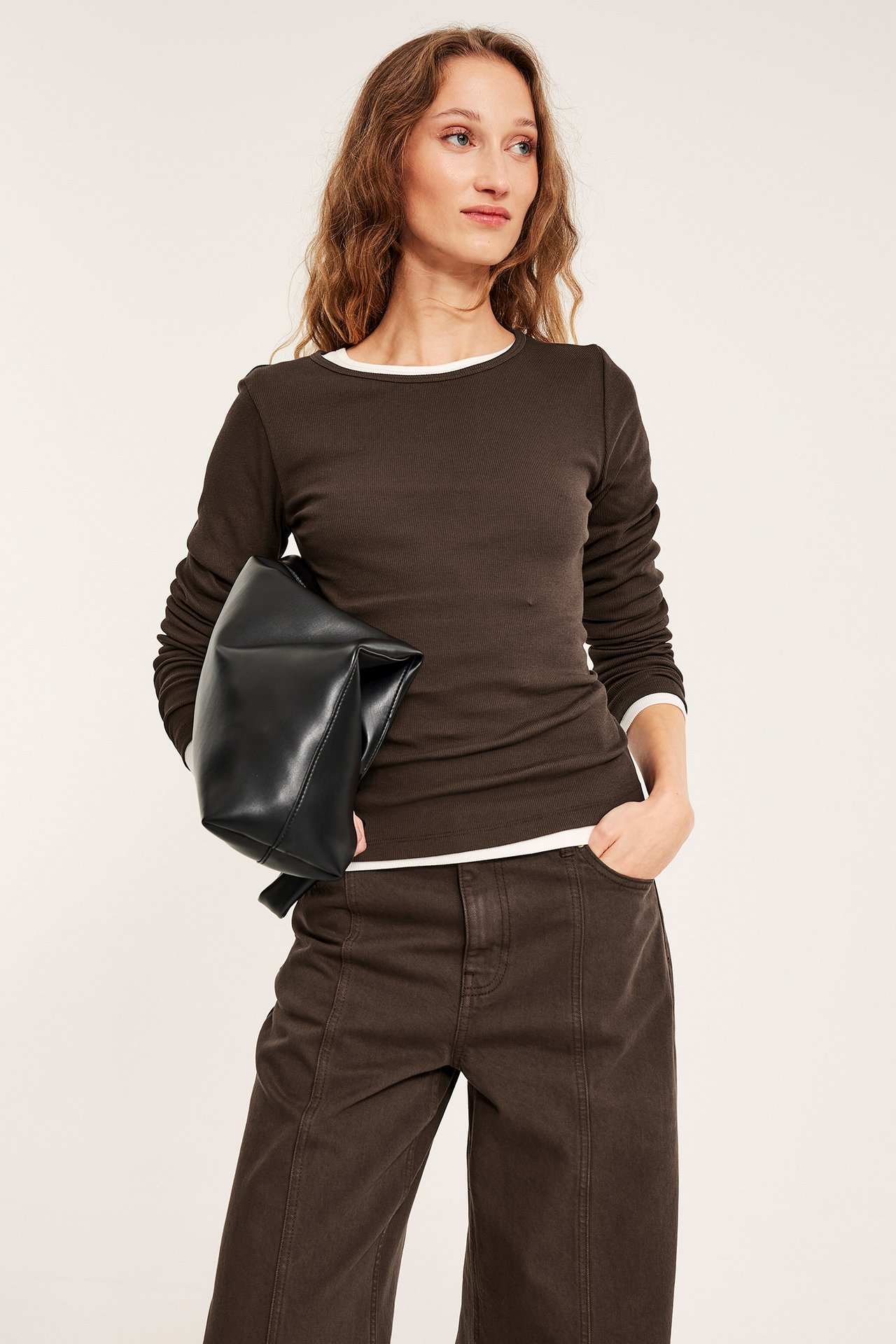 Widok z przodu: Kobieta w ciemnobrązowym prążkowanym topie z białym obszyciem, brązowe spodnie, trzyma czarną torbę.