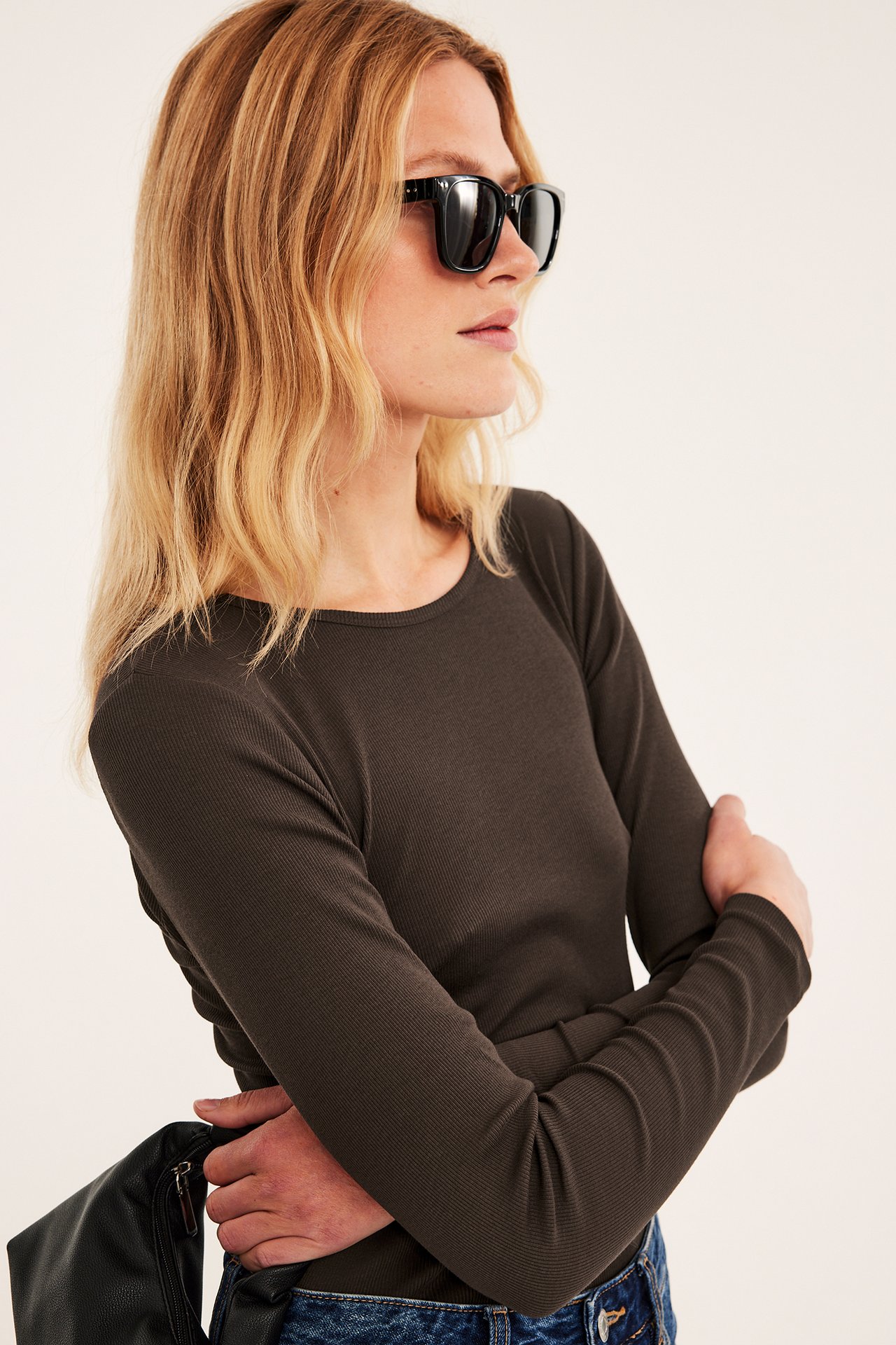 Kvinne i mørk oliven ribbet topp, blå jeans, svarte solbriller, holder svart mageveske.