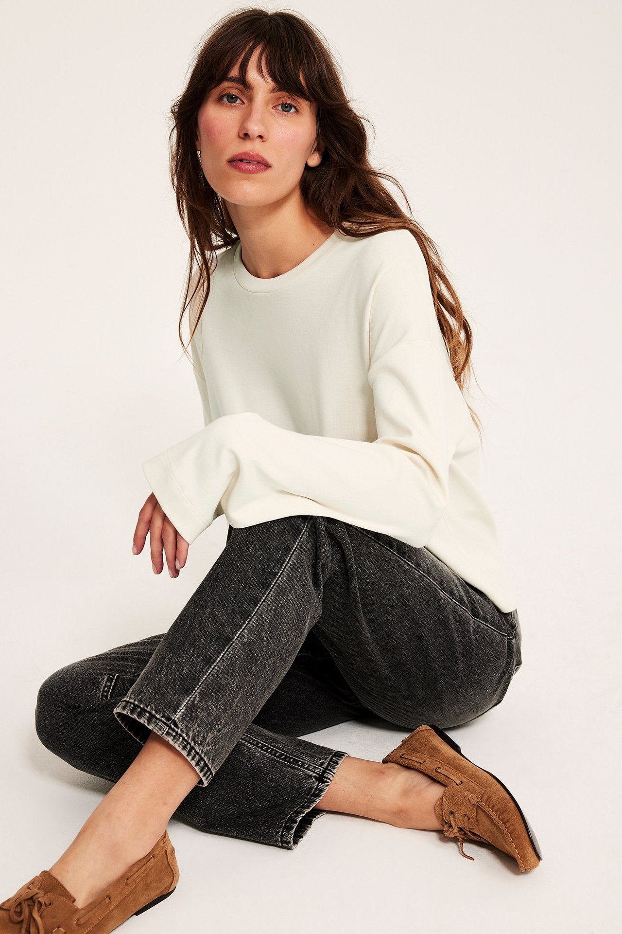 Widok z przodu: kobieta w luźnym, kremowym bawełnianym T-shircie z długim rękawem, ciemnoszarych jeansach i brązowych mokasynach.