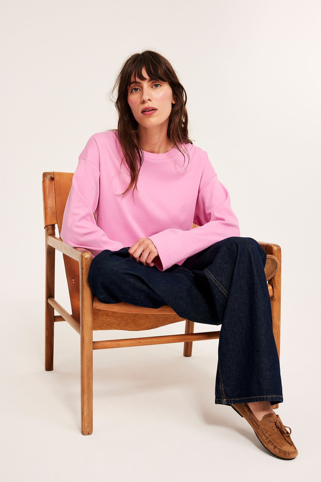 Widok z przodu: kobieta w luźnej różowej bawełnianej koszulce z długim rękawem, ciemnoniebieskich dżinsach i brązowych mokasynach.