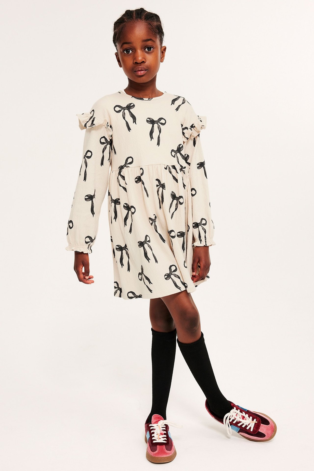 Framifrån: Barn i ljusbeige jerseyklänning med svarta rosetter, svarta strumpor och vinröda sneakers.