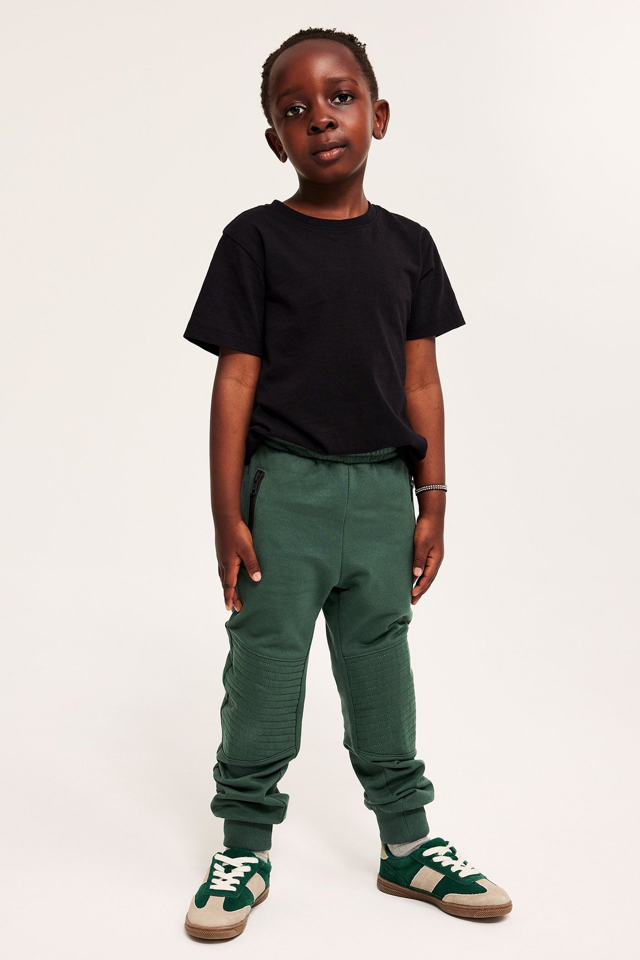 Edestä: Poika vihreissä puuvillacollegehousuissa vahvistetuilla polvilla, musta t-paita ja lenkkarit.