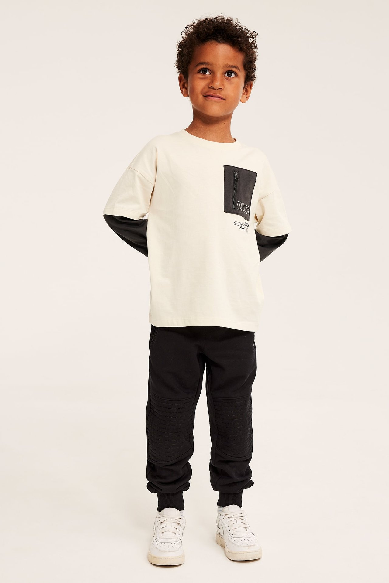 Front view: Kid i skiktad benvit t-shirt med mörk ficka med dragkedja, svarta joggers, vita sneakers.