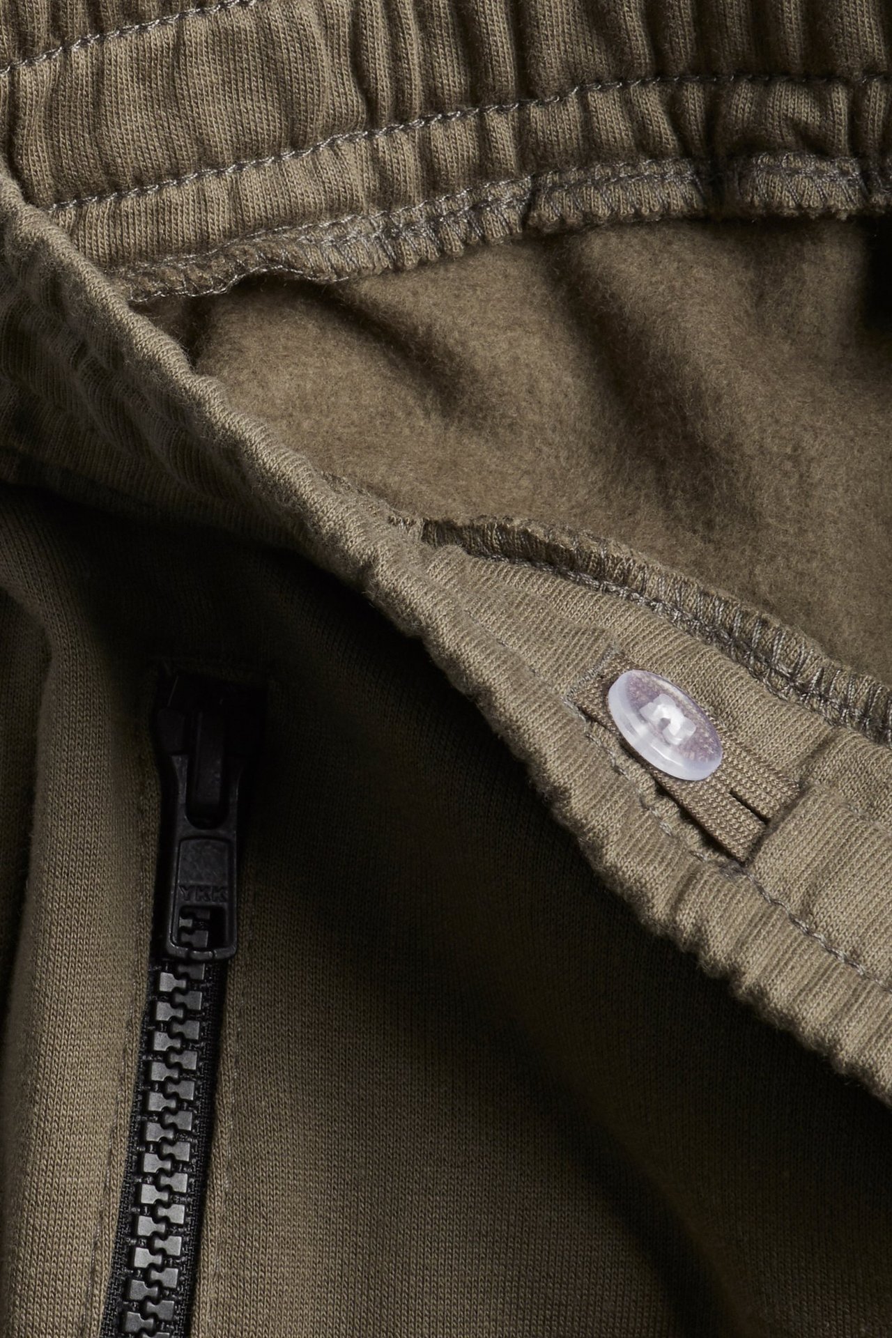 Lähikuva ruskeista puuvillaisista collegehousuista, joissa joustava vyötärö, musta vetoketju ja läpinäkyvä nappi.