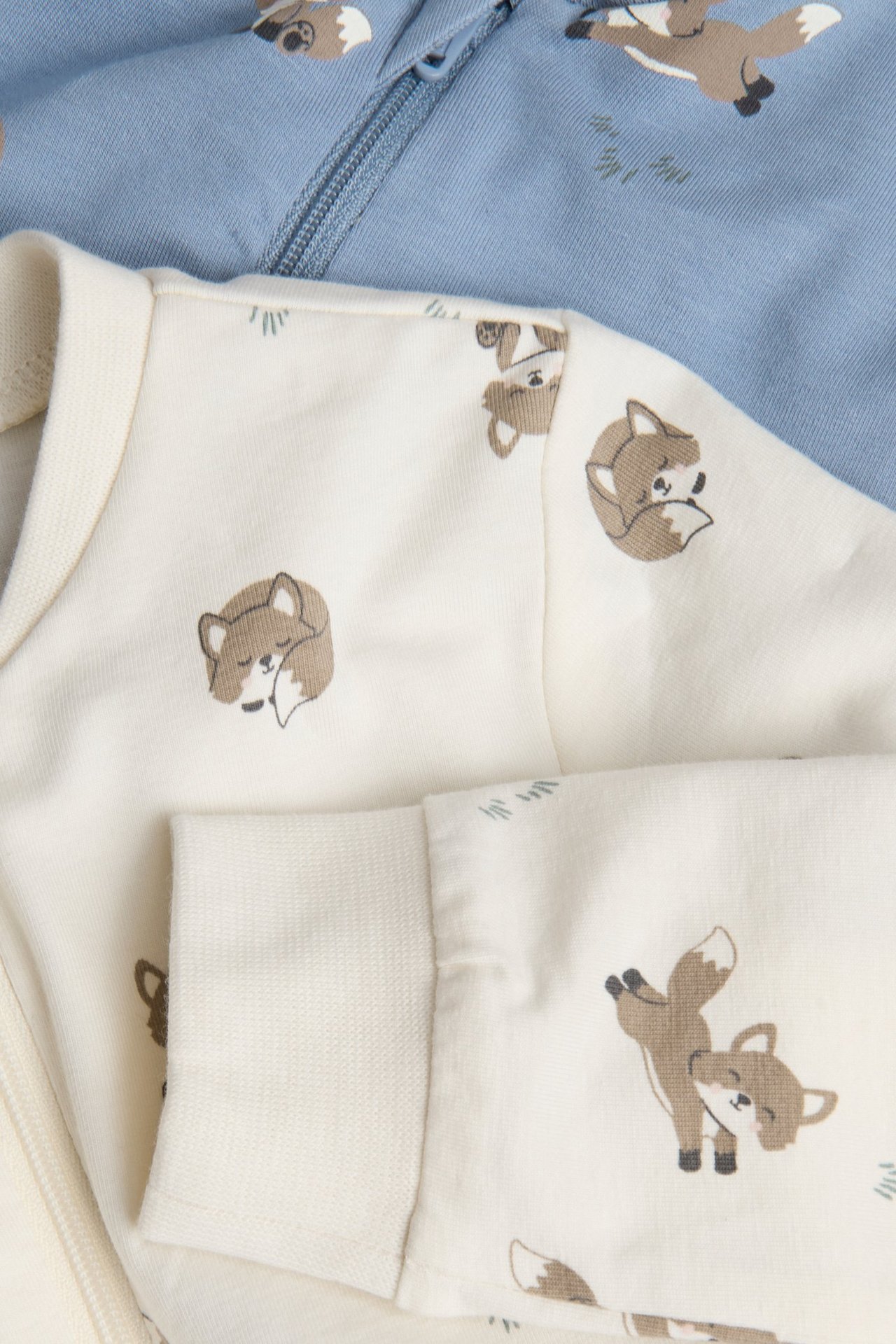 Zbliżenie na kremowy pajacyk niemowlęcy w lisy z zamkiem, z niebieskim pajacykiem w lisy z tyłu.